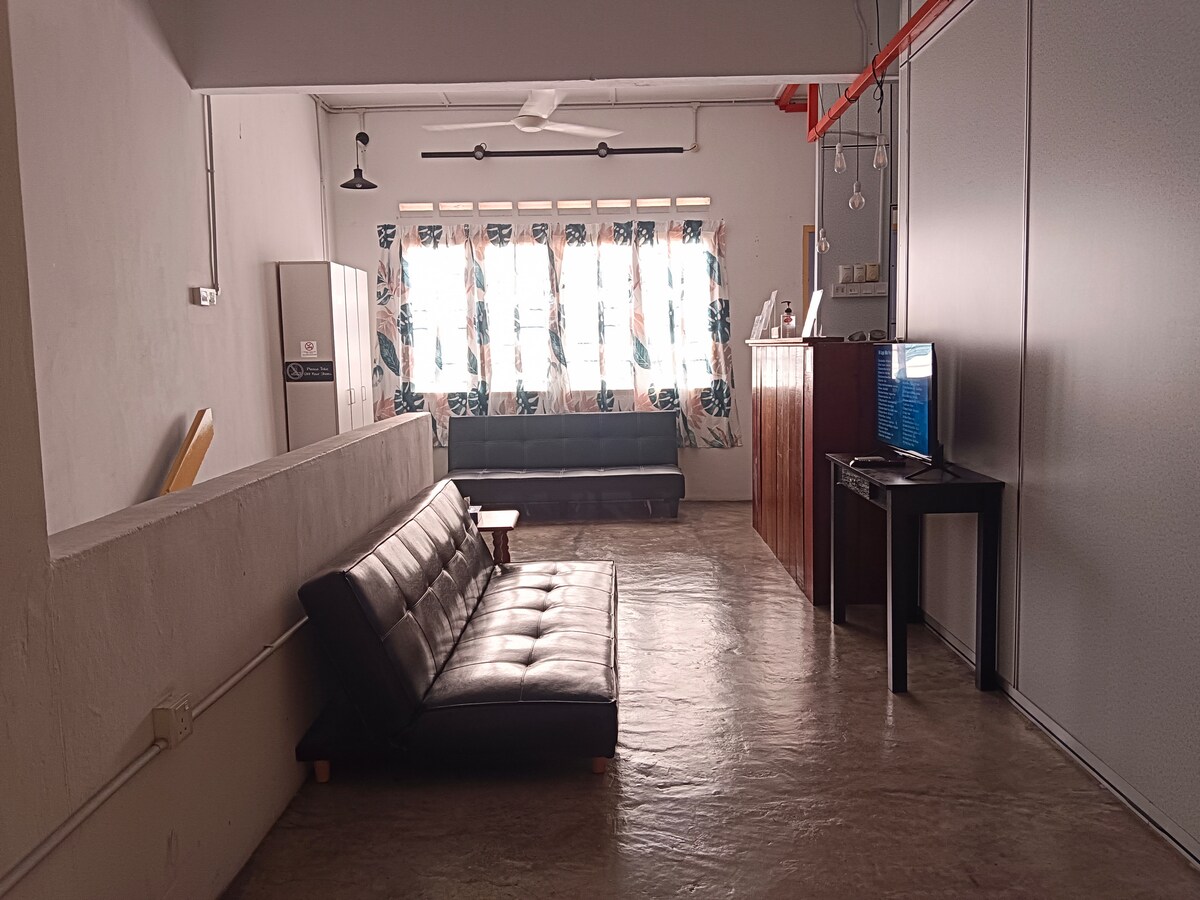 A shared common area features a dark sofa and a sleek black TV stand. Natural light filters through large windows adorned with floral curtains. A minimalist, contemporary feel is created by the polished concrete flooring and white walls, complemented by a ceiling fan and lighting fixtures.