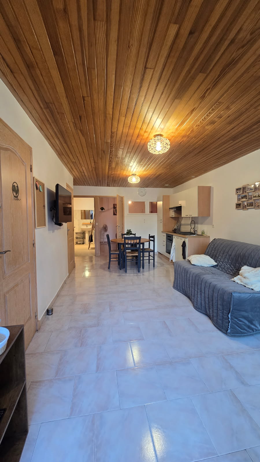 The living space features a wooden ceiling enhancing the warm ambiance. A sofa is positioned beside a dining table with four chairs. Adjoining kitchen elements are visible, providing functionality. Light-colored floor tiles contribute to the overall brightness of the room.