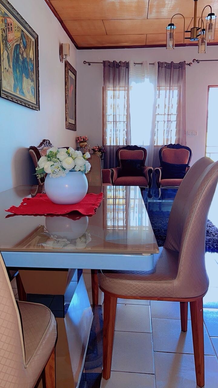 A dining area is presented with a glass table and six beige chairs. A floral arrangement in a white vase sits centrally on the table. Two armchairs with a rich fabric are positioned nearby, and light filters through sheer curtains, creating a bright and inviting space.
