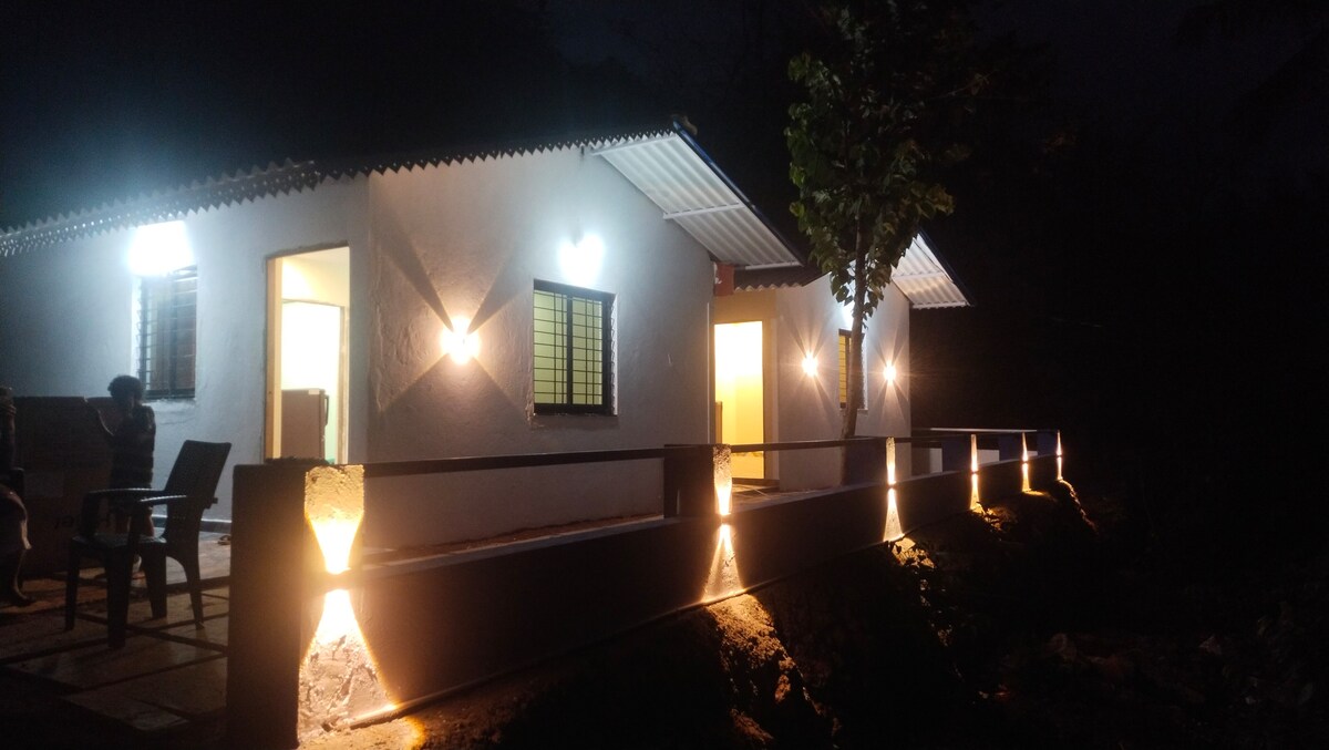 The exterior of a cozy, well-lit home is showcased at night. Soft lighting highlights the facade and pathway, accompanied by outdoor seating. A tree stands nearby, contributing to the peaceful ambiance of the setting.