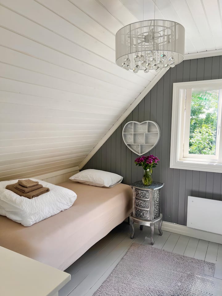 Chambre charmante avec plafond en pente et lit simple. La lumière naturelle de la fenêtre et les surfaces claires créent une atmosphère calme et agréable.