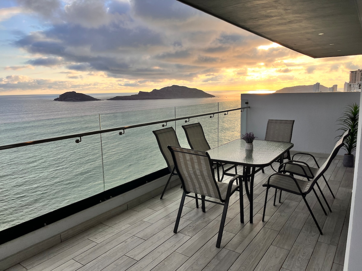 A private balcony features a dining table and several chairs, overlooking the serene Pacific Ocean and islands in the distance. The warm hues of the sunset reflect on the water, and the space is designed for relaxation and scenic views of the surrounding landscape.