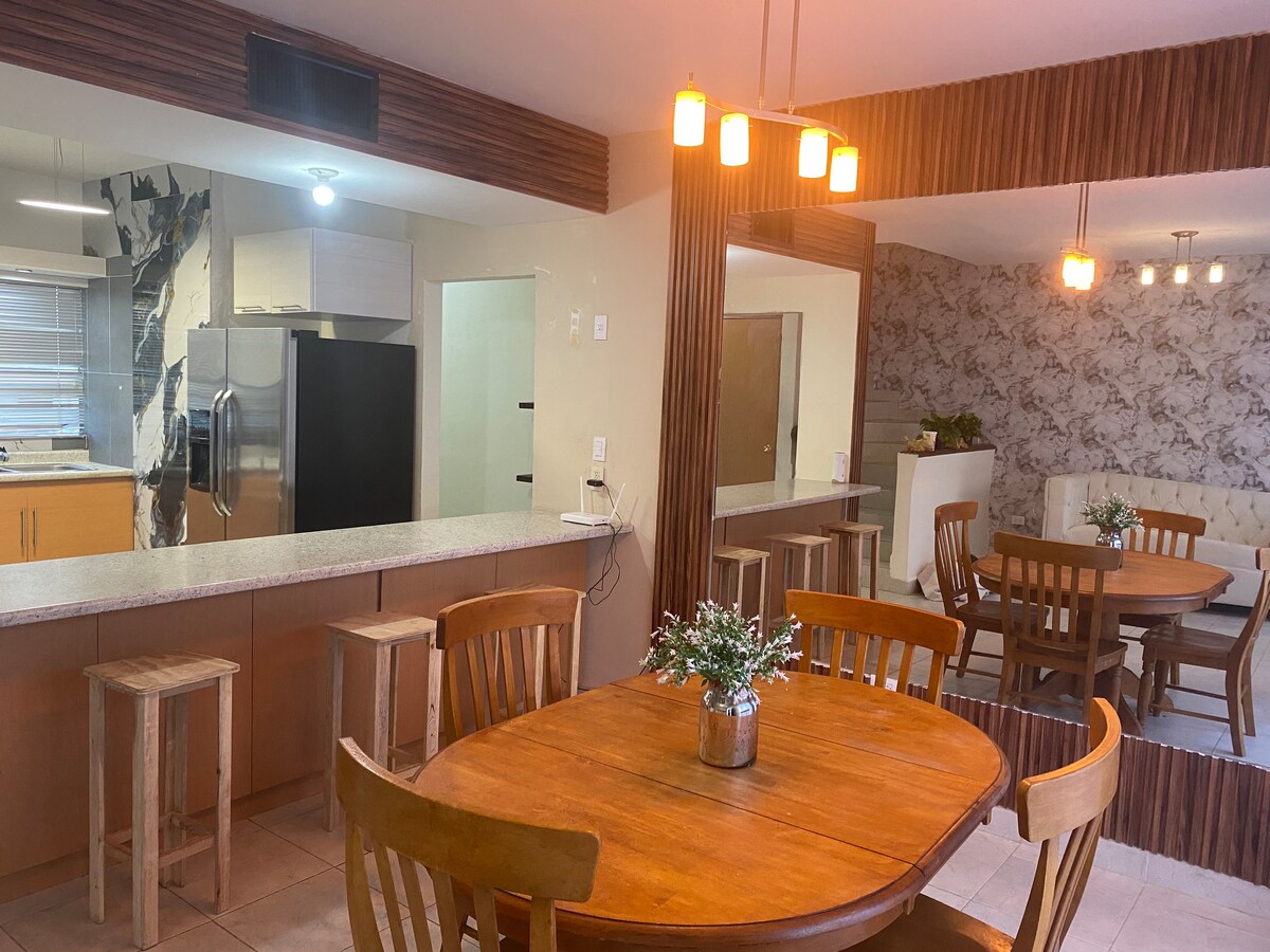 The image shows a dining area with wooden tables and chairs. One round table is set for four, while a larger dining area features a rectangular table. A kitchen counter is visible, equipped with modern appliances. Decorative light fixtures provide overhead illumination.