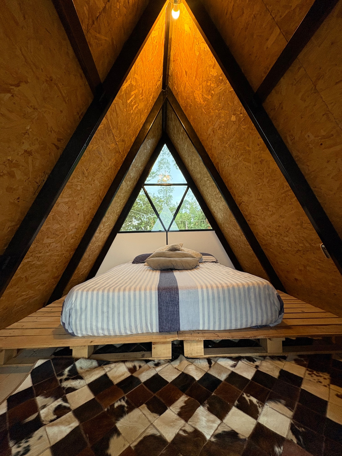 An A-frame structure features a spacious bed with a striped blanket, positioned on a wooden platform. A large window with a diamond design allows natural light to fill the room, revealing the outdoor greenery through the openings in the wooden walls.