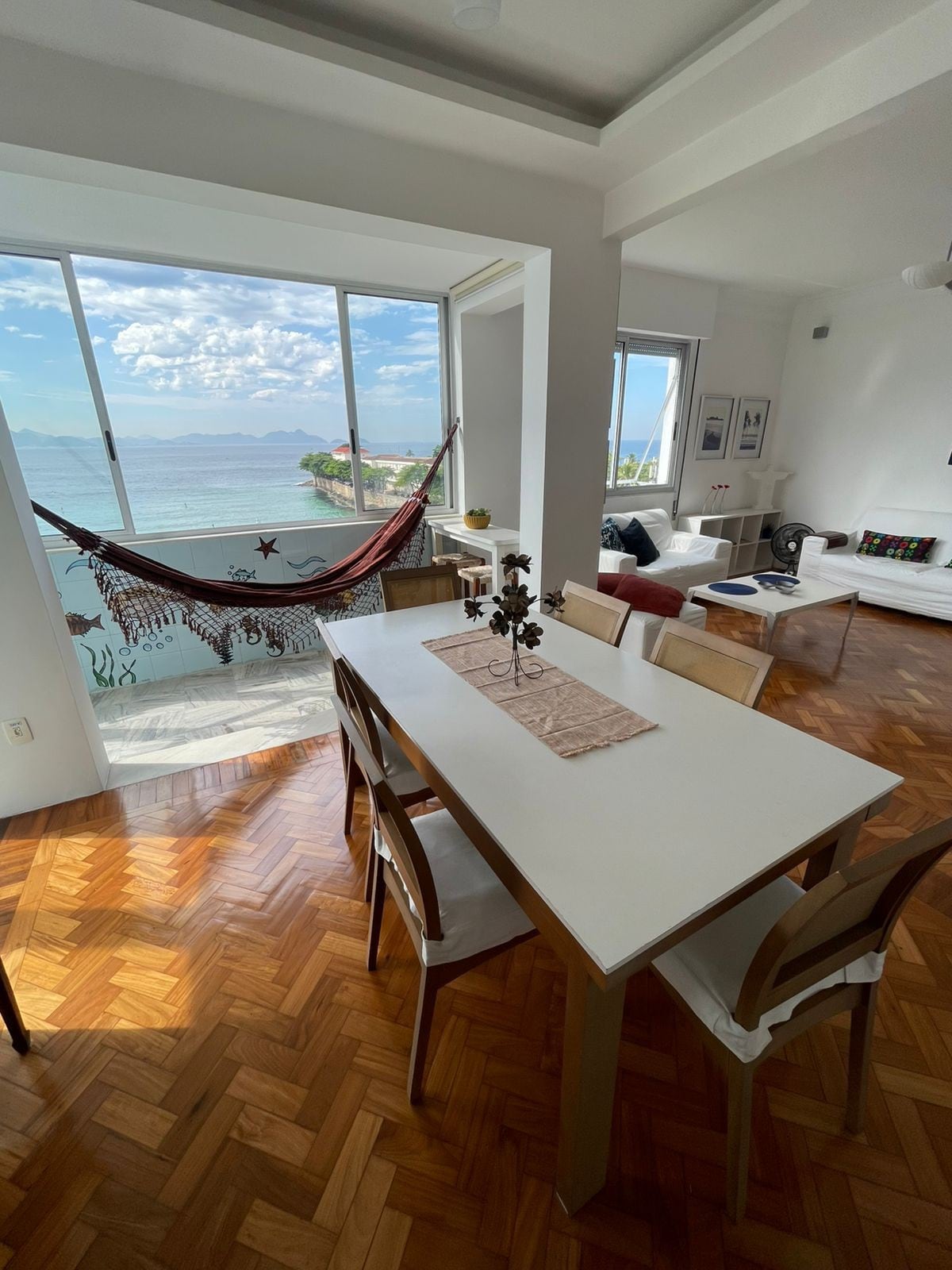A bright dining area is illuminated by natural light streaming through large windows. A stylish wooden table with several chairs is positioned near the window, overlooking scenic ocean views. A hammock hangs invitingly to the side, enhancing the relaxing atmosphere of the space.