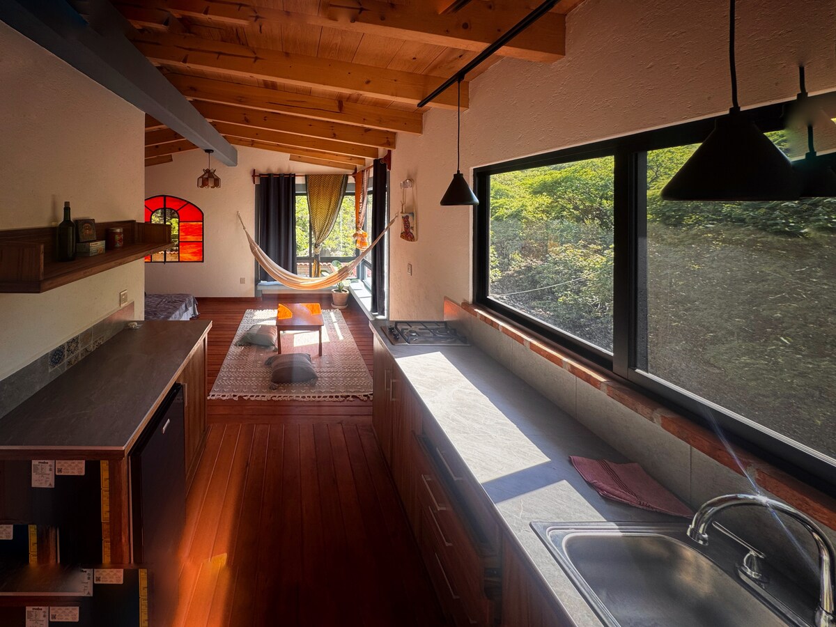 A spacious kitchen featuring wooden cabinetry and a sleek countertop is illuminated by natural light from large windows. A hammock is visible in the adjacent living area, complemented by a colorful rug. Exposed wooden beams add warmth to the contemporary design.