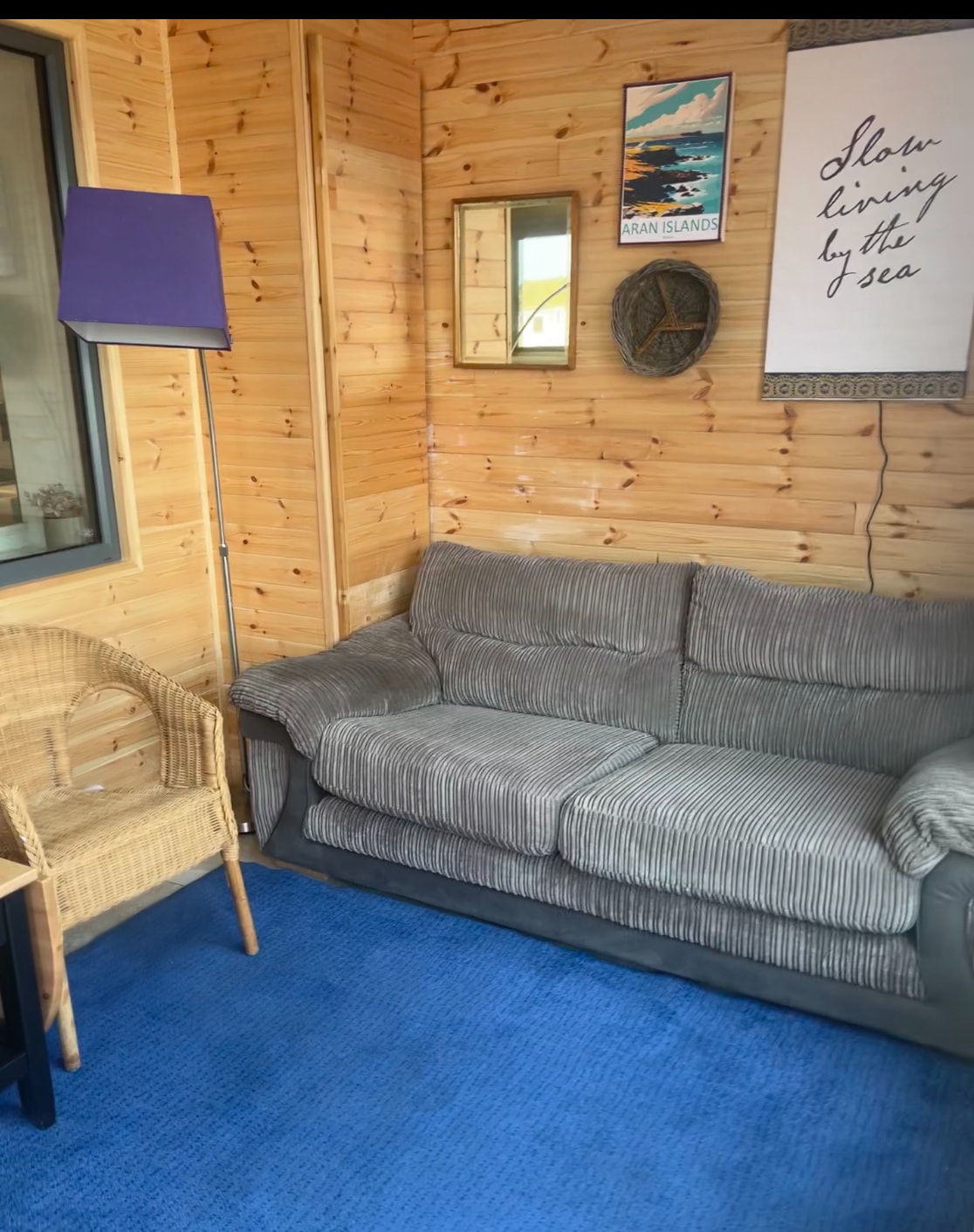 A comfortable sitting area is presented with a plush gray sofa and a woven chair. Light wood paneling adorns the walls, complemented by a soft blue carpet. A reflective mirror and a wall art piece with a coastal theme enhance the space.