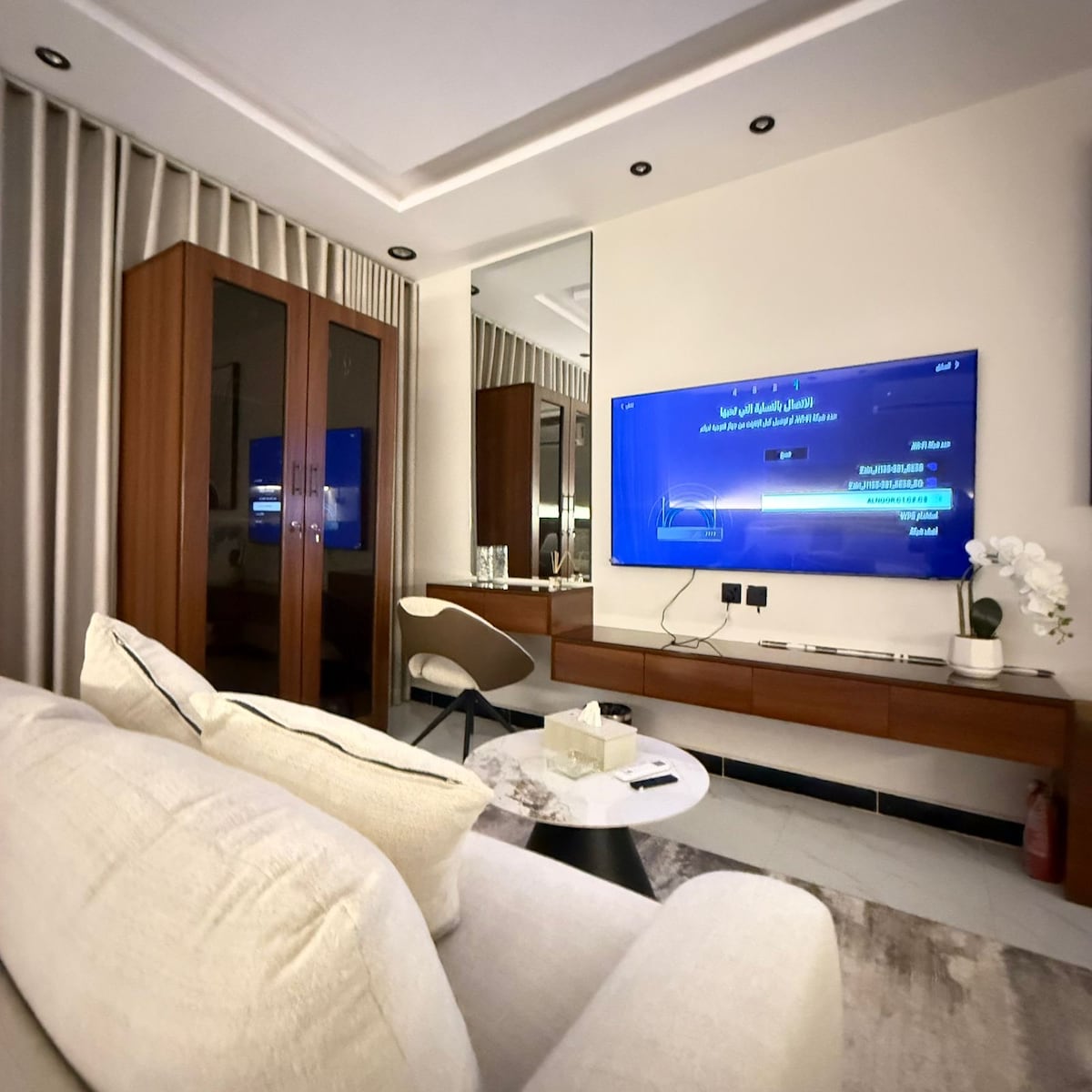 A cozy seating area is presented with a spacious sofa, facing a large TV mounted on the wall. A stylish mirror reflects the room's elements, while a sleek cabinet provides storage. Neutral tones and soft lighting create a calm environment.