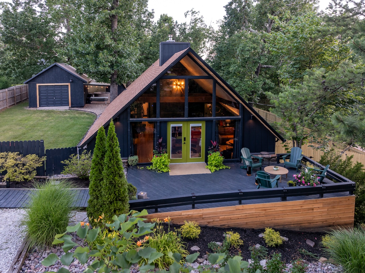 An A-frame house is set within a lush landscape, framed by tall trees. A spacious deck features seating for relaxation, with a small table surrounded by green chairs. The vibrant entrance showcases large glass doors, leading to the inviting interior. A detached garage is visible in the background.