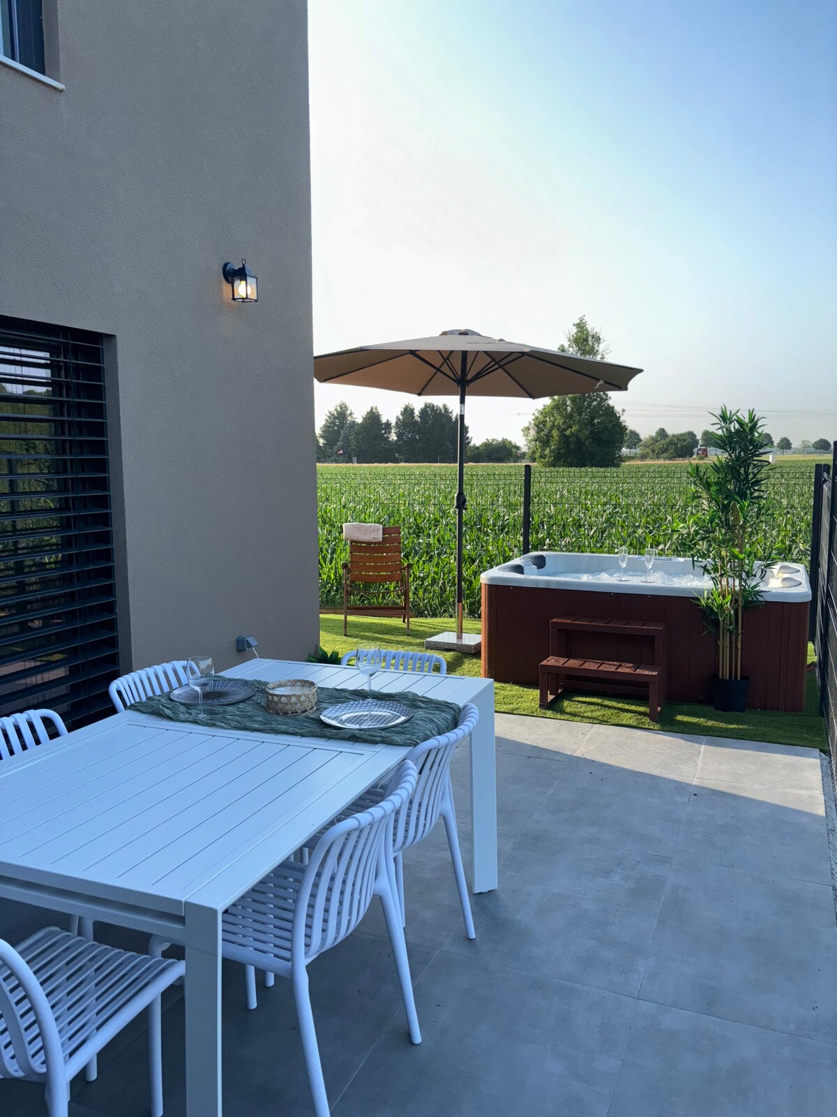 An outdoor terrace is shown, featuring a dining table with white chairs and a parasol providing shade. In the background, a jacuzzi is nestled in a private space surrounded by greenery. A scenic view of a field enhances the relaxing atmosphere.