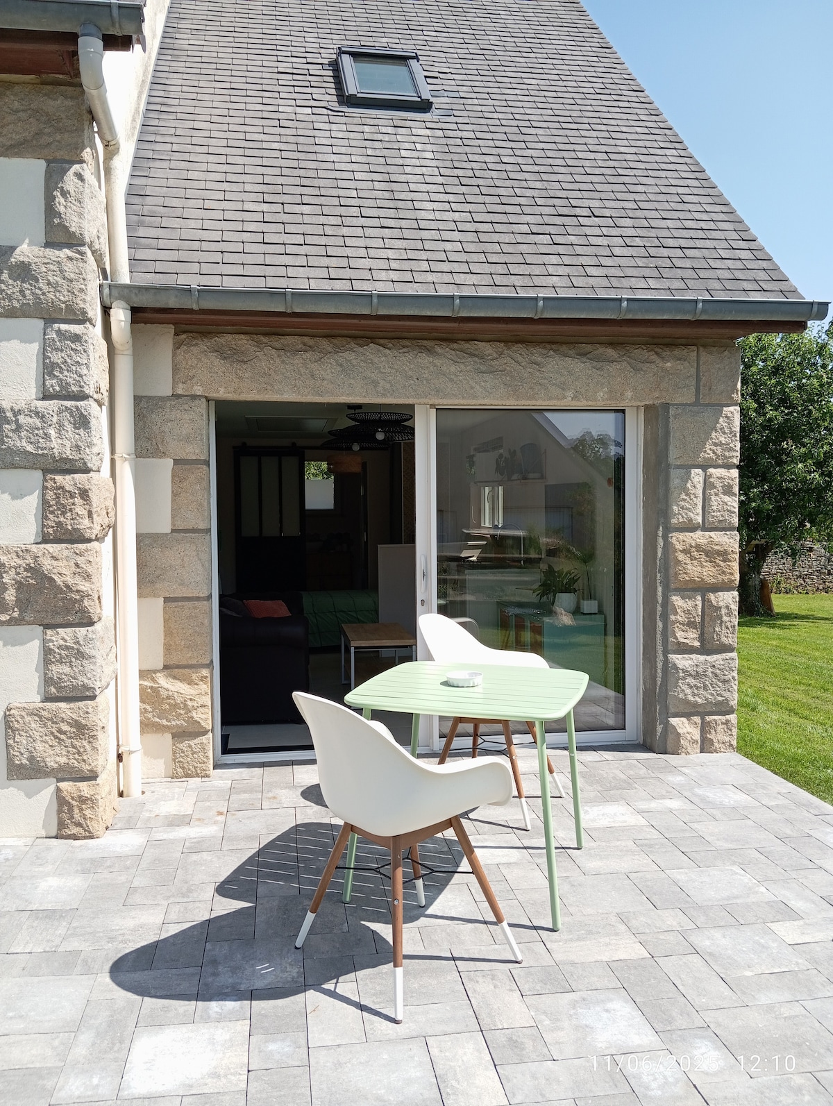 A terrace features a light green table accompanied by two white chairs, set on a paved stone surface. Sliding glass doors provide access to the interior space, with a view of the garden visible beyond.