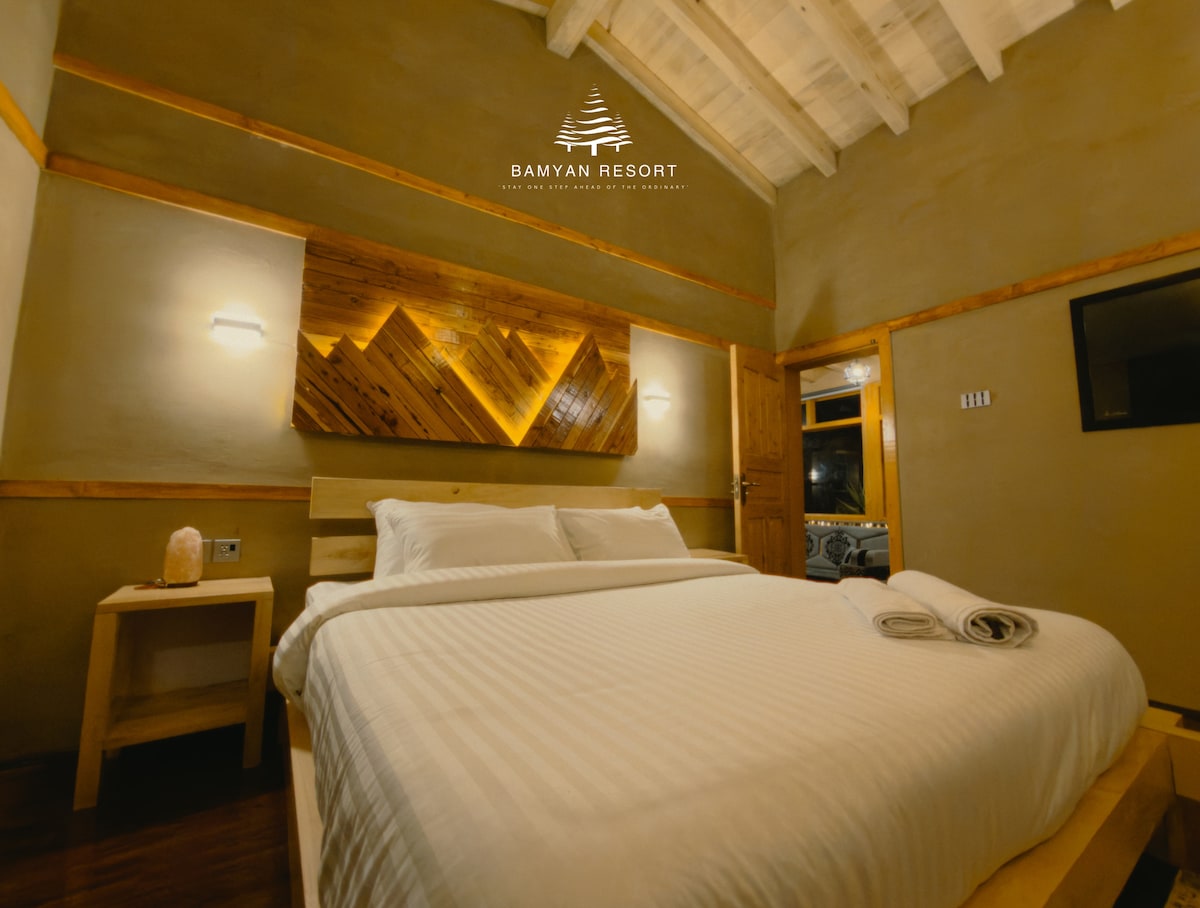 A spacious sleeping area features a light-colored bedspread draped over the large bed, complemented by soft towels neatly rolled on the side. The wooden accent wall behind the bed adds warmth, while gentle lighting enhances the serene ambiance of the room.