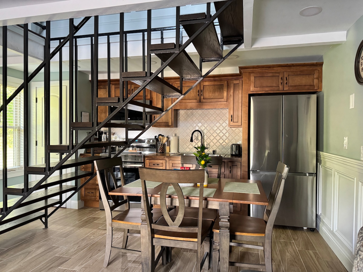 A modern kitchen is visible, featuring wooden cabinetry and stainless steel appliances. A dining table with four chairs is positioned in the foreground, while a metal staircase leads to the upper level. Natural light floods the space through large windows.