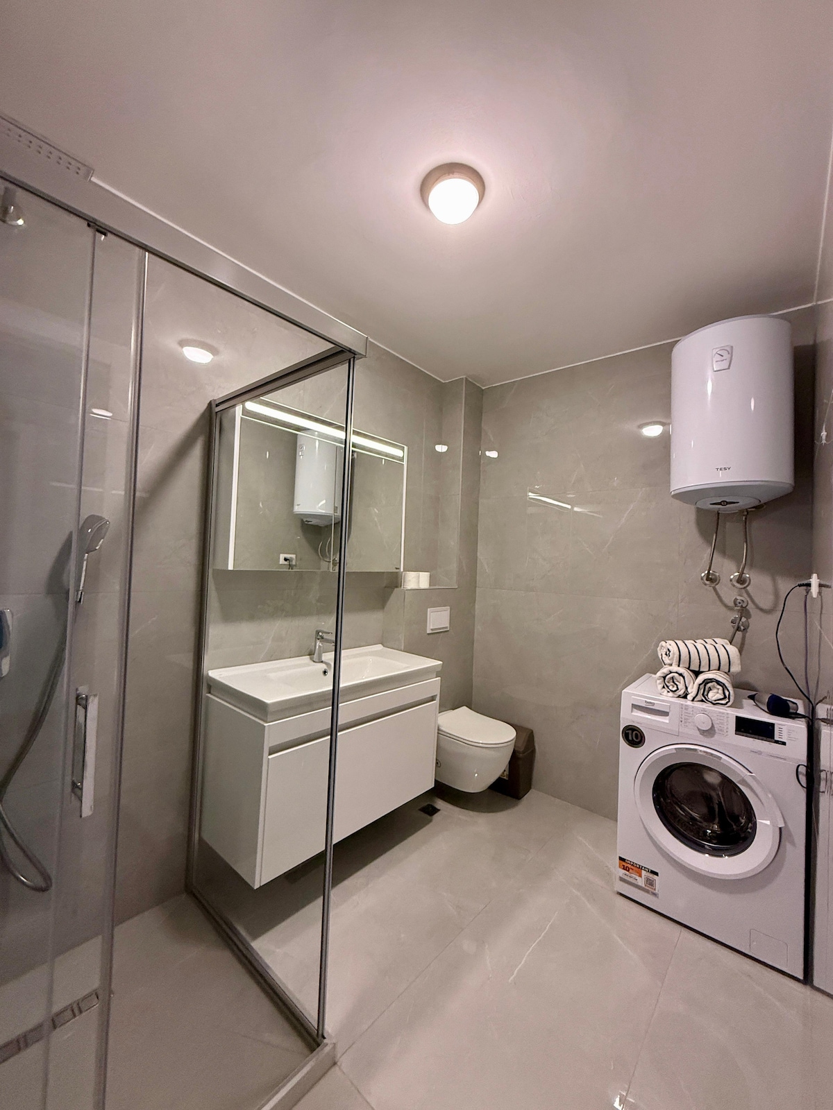 A modern bathroom is presented with sleek gray tiles and a spacious walk-in shower enclosed in glass. A white vanity with a mirror is located above a contemporary sink, alongside a washing machine and a compact toilet, providing functionality in a clean and unadorned space.