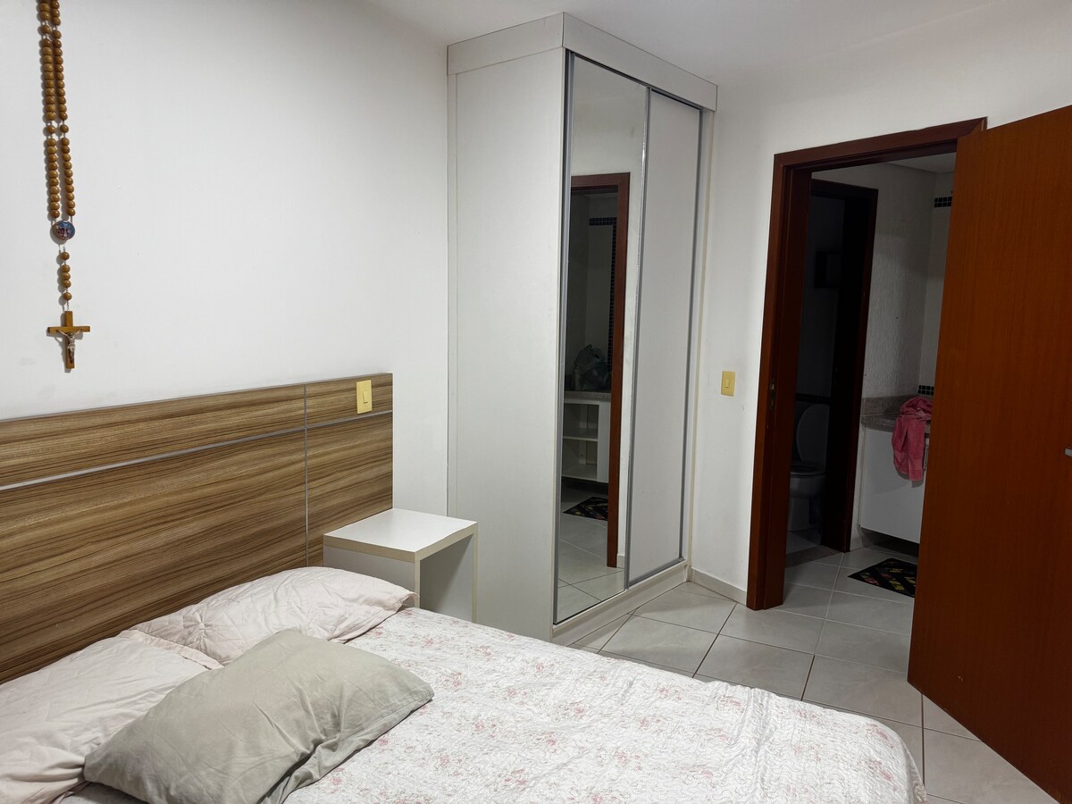 A cozy bedroom features a spacious bed adorned with light-colored bedding. A mirrored wardrobe reflects the room's design, while a small nightstand sits beside the bed. A crucifix hangs on the wall, adding a touch of character. The doorway leads to a bathroom.