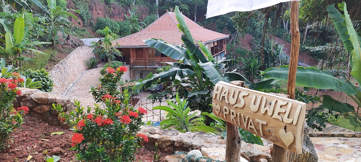 A wooden sign labeled 'Haus Uweli Privat' is prominently displayed among lush green foliage and colorful flowering plants. In the background, a traditional wooden house is nestled on a hillside, surrounded by a tranquil natural setting.