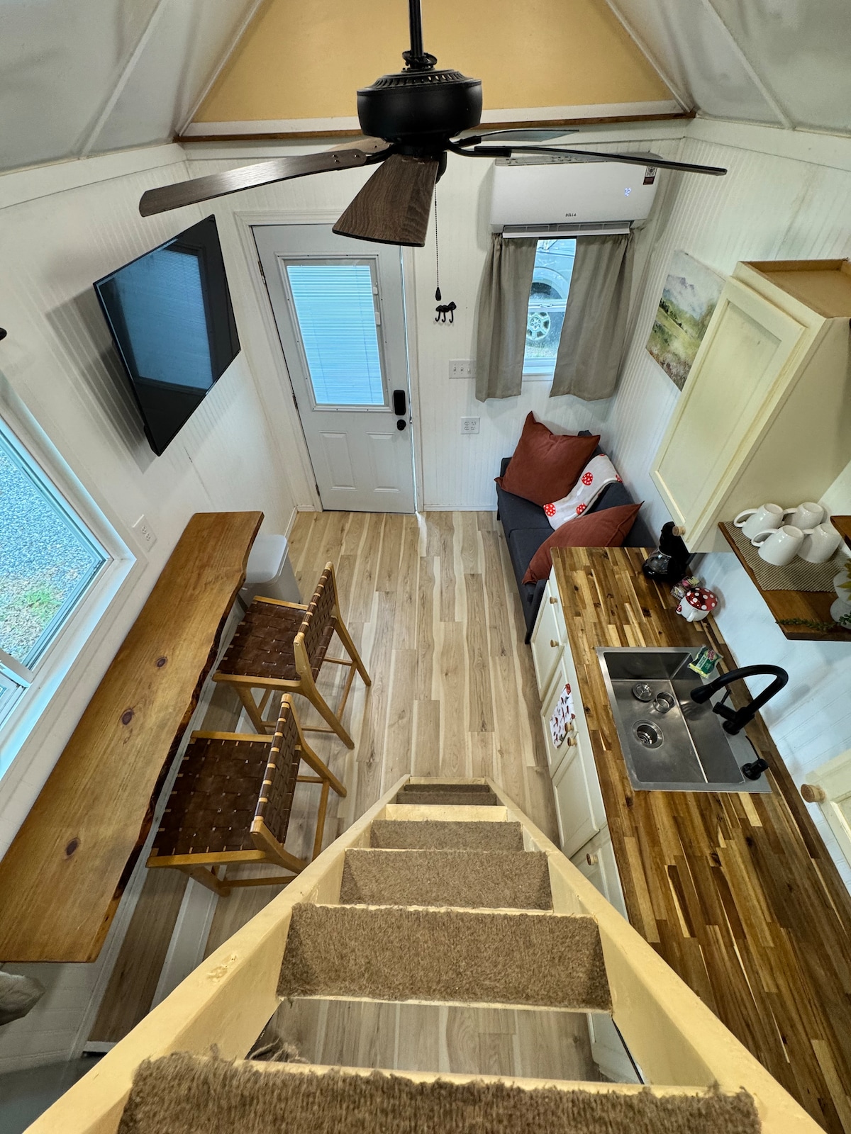 A view from above showcases the interior layout, featuring a staircase leading to the loft. The kitchenette is equipped with a sink and modern appliances. A cozy living space includes a small sofa and a dining bar with two stools. Natural light illuminates the area.