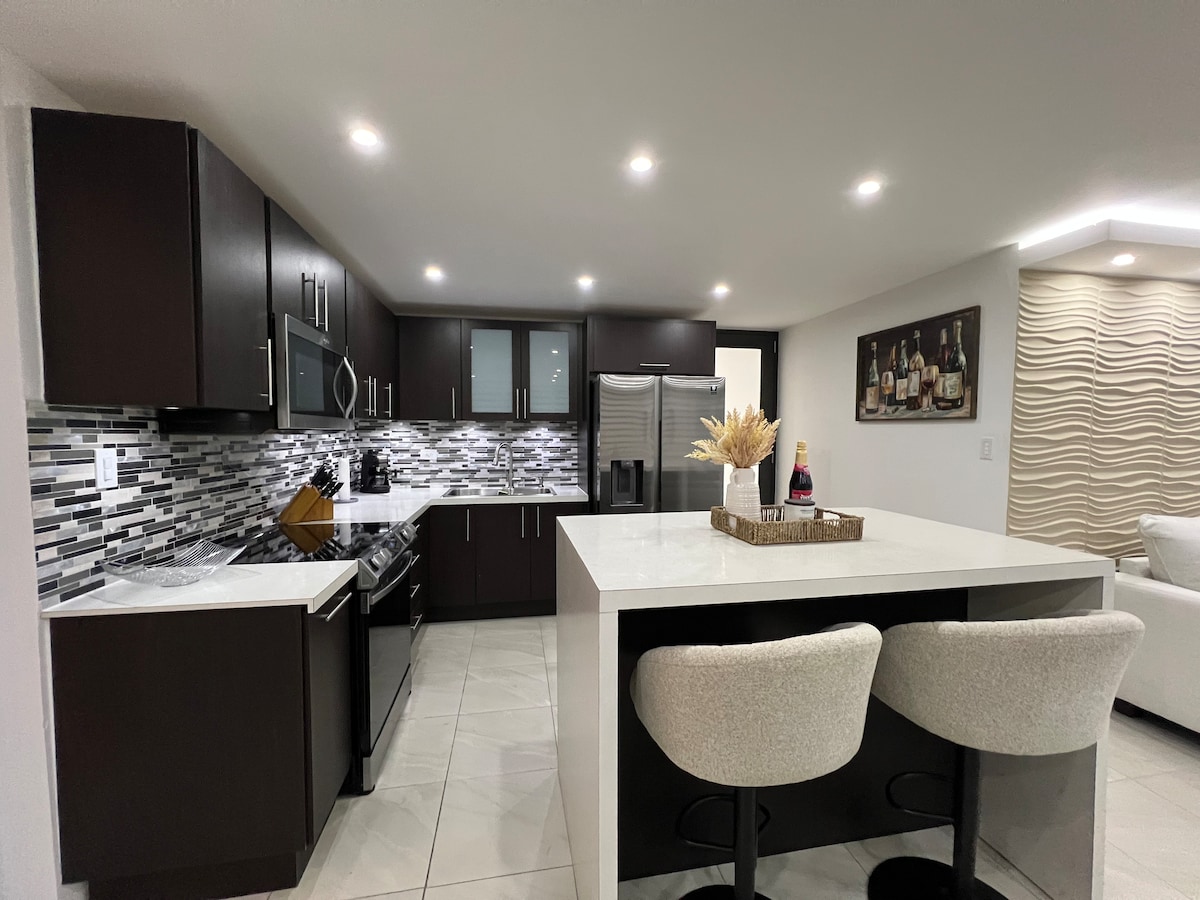 A modern kitchen is showcased with sleek dark cabinetry and a striking backsplash of gray and white tiles. Stainless steel appliances are included, complemented by a spacious white countertop. Two bar stools are positioned at a breakfast bar, enhancing the kitchen's functionality.
