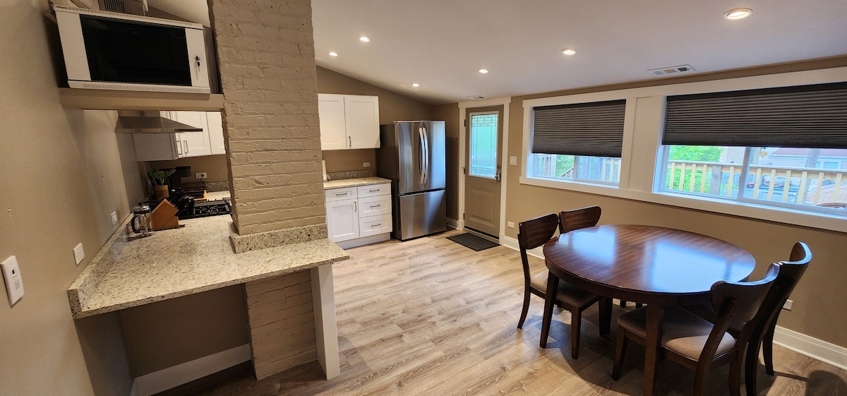 A modern kitchen and dining area is presented, featuring a round wooden table with four chairs. New appliances, including a refrigerator and microwave, are visible. Light-colored cabinets complement the granite countertop, while natural light enters through the window with a view of the outdoor space.
