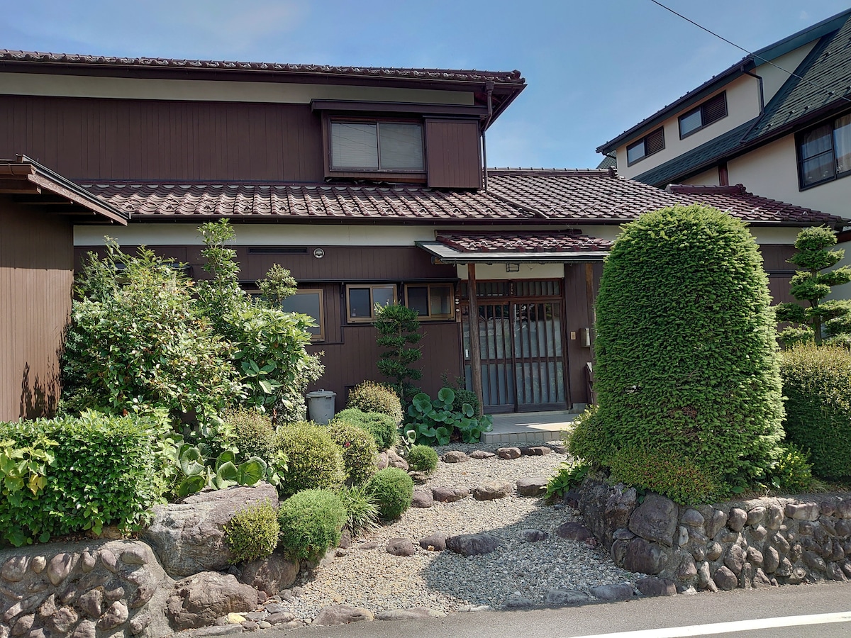 A traditional house with a brown wooden exterior is surrounded by well-maintained greenery. A gravel pathway leads to the entrance, flanked by decorative shrubs and small trees. The roof features dark tiles, and a fence can be seen in the background.