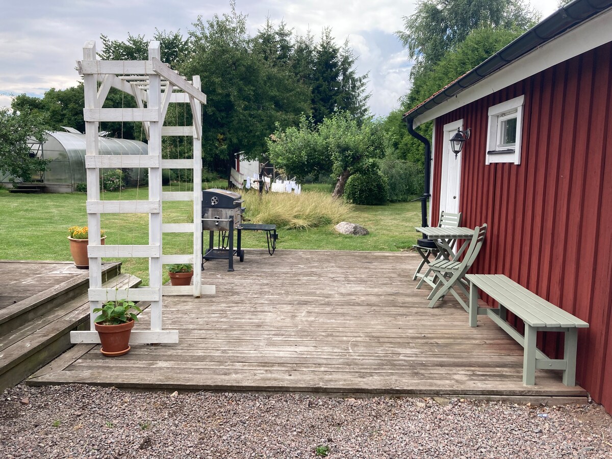 An outdoor patio area is featured, with a wooden deck extending from the side of a red building. A small table and two benches are present alongside potted plants. A grill is positioned nearby, with a trellis adding structure to the space.