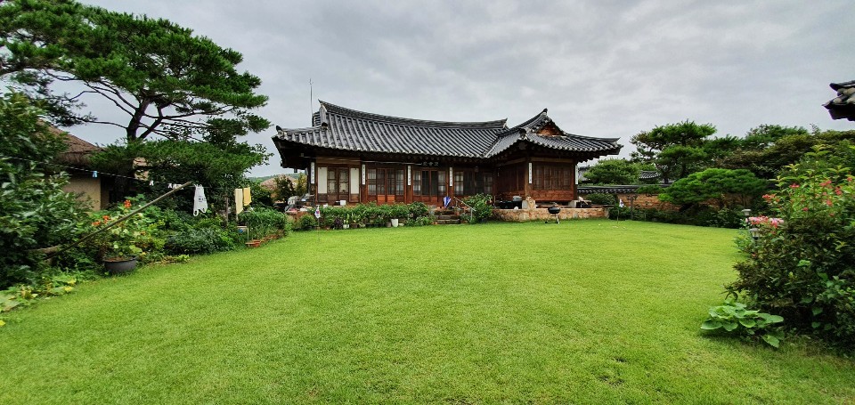 A traditional Korean house is set amidst a well-maintained garden featuring lush grass and various plants. The building's wooden exterior is complemented by a gracefully curved roof. Surrounding trees provide a natural backdrop, enhancing the serene environment.