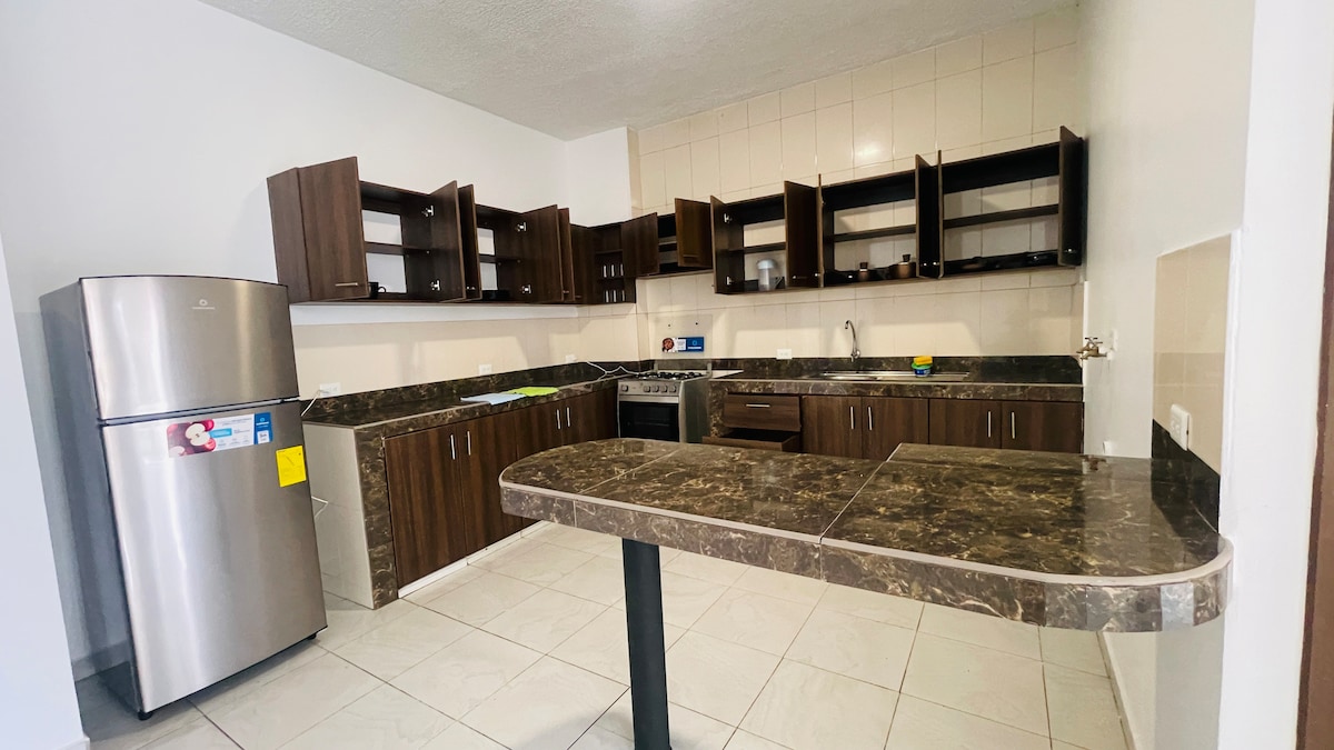 The kitchen features wooden cabinetry with upper and lower storage, and is equipped with a stainless steel refrigerator and a gas stove. A multi-purpose countertop provides additional workspace, while tile flooring enhances the overall modern feel of the space.