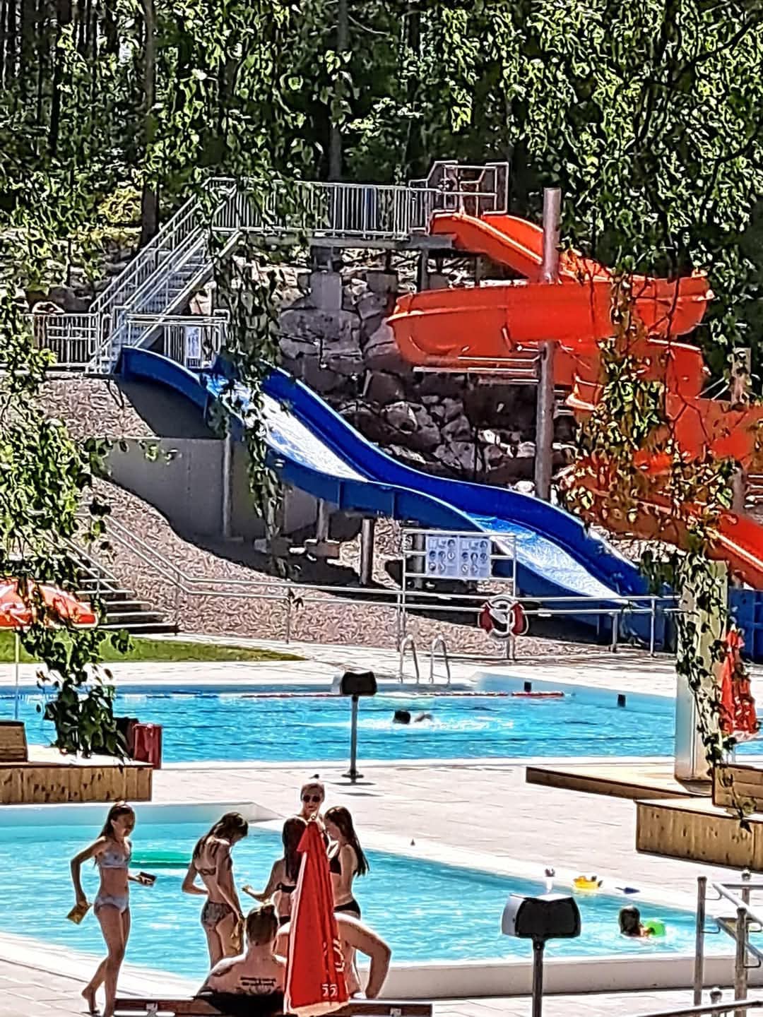 A children's aquatic area is visible, featuring a blue and orange water slide positioned above a pool. Several individuals are enjoying the water, while lounge chairs and vibrant umbrellas provide shaded seating nearby. The surrounding greenery adds to the setting.