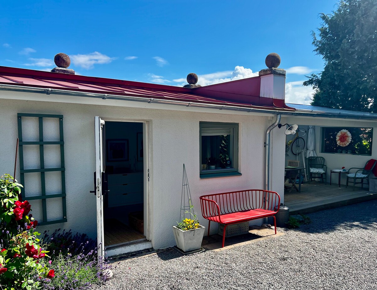A white exterior is complemented by a red metal roof. The entrance features a simple wooden door, with a cozy red bench placed outside. Flower pots are positioned near the entrance, adding a touch of greenery. A shaded patio area is visible to the right.