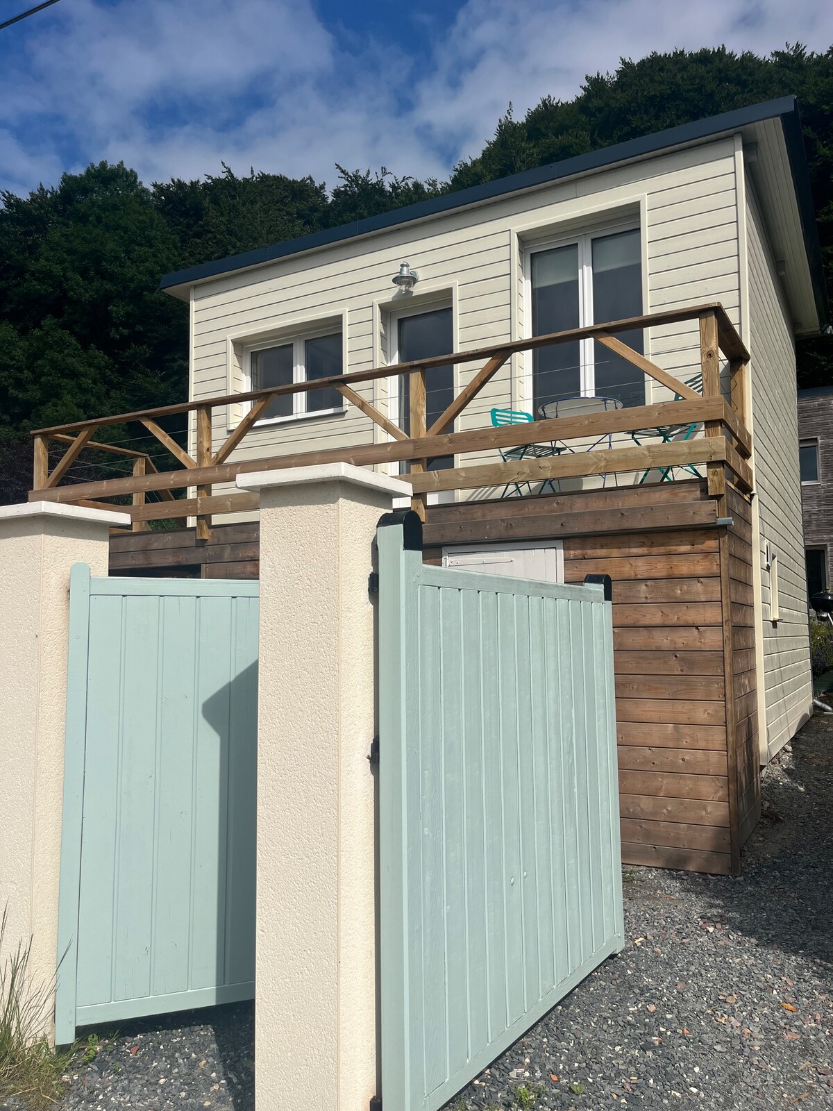 A charming little annex house is presented with a wooden terrace overlooking nature. The exterior features light-colored siding contrasted with wood detailing, while a gated entrance leads to the property. Large windows invite natural light into the living space.