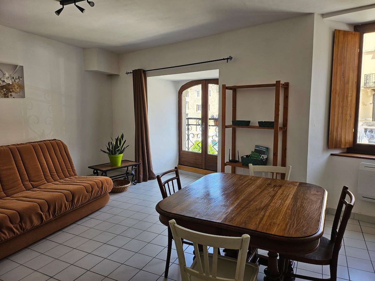 A bright living area features a spacious convertible sofa and a wooden dining table accompanied by four chairs. Natural light enters through a window with wooden shutters, highlighting the greenery of a potted plant. A shelf displays green decor, contributing to a homey atmosphere.
