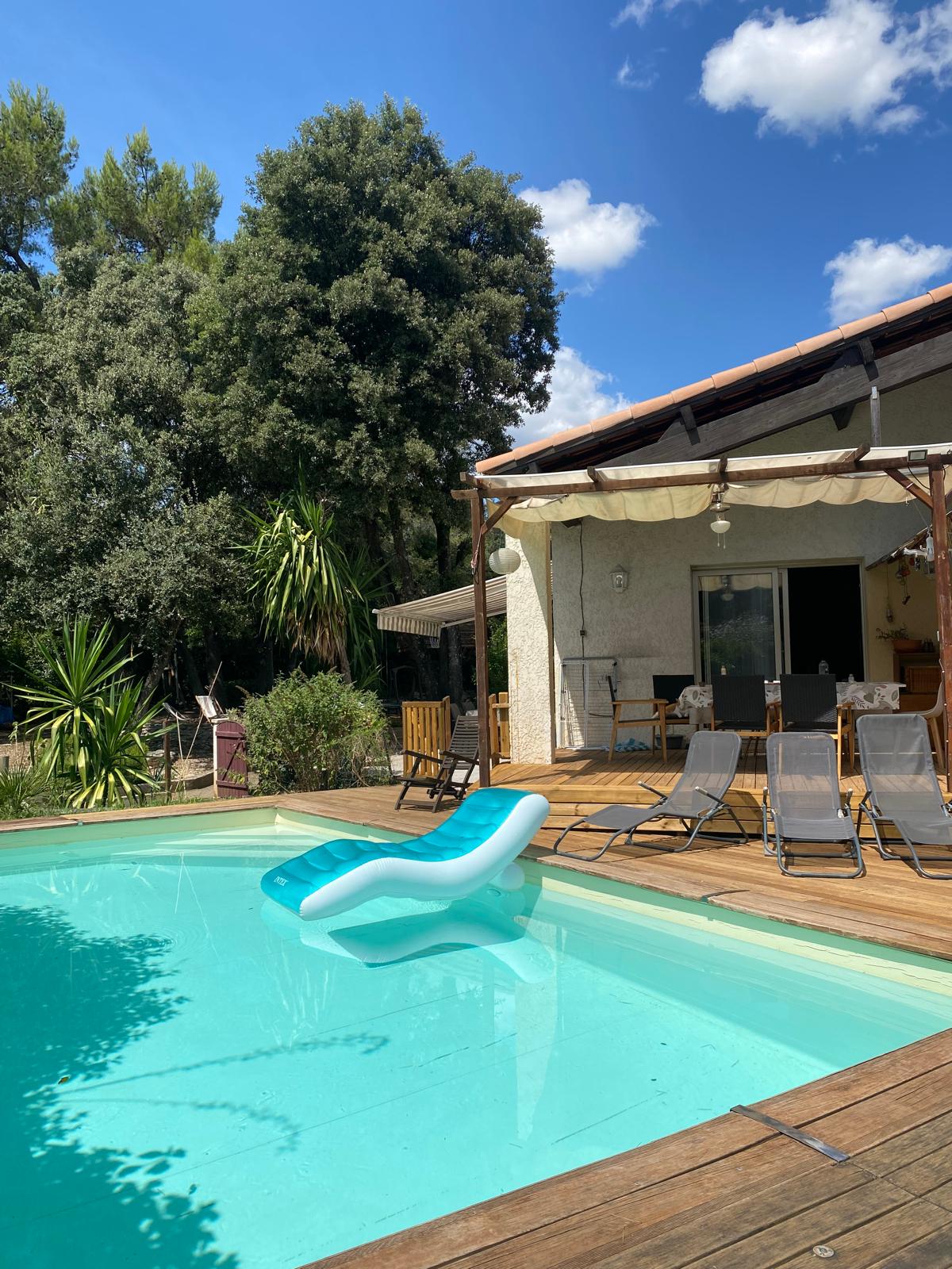 A swimming pool is framed by wooden decking, with a large green slide positioned on one side. Sun loungers are arranged nearby, and lush greenery surrounds the area, creating a tranquil outdoor setting. The house features a covered patio visible in the background.