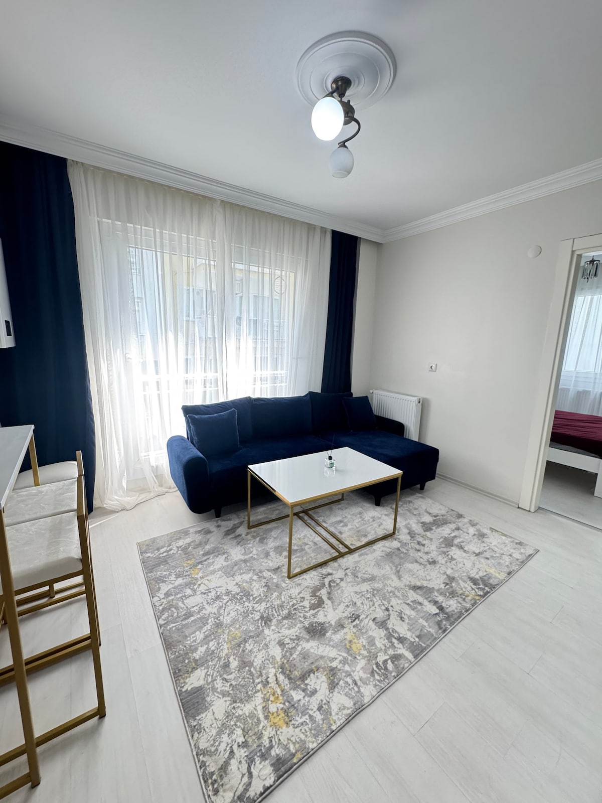 A living area features a navy blue sofa positioned beside a glass coffee table on a patterned rug. Natural light streams through sheer curtains covering a large window, complementing the minimalist decor. A small dining area with gold accents is visible in the background.