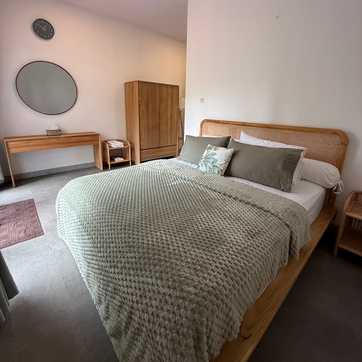 A well-appointed bedroom features a spacious bed with neutral-toned bedding, accompanied by a textured throw blanket. Natural light fills the room, which includes a wooden wardrobe, a console table, and a round mirror. Soft accents are evident in the overall design.