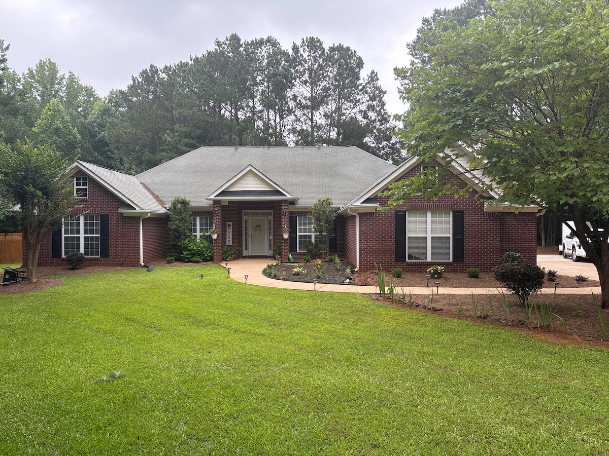 An inviting single-story home is surrounded by lush green lawns and neatly landscaped gardens. The brick exterior features large windows and a central entrance framed by carefully tended shrubs and trees, creating a pleasant and welcoming appearance.