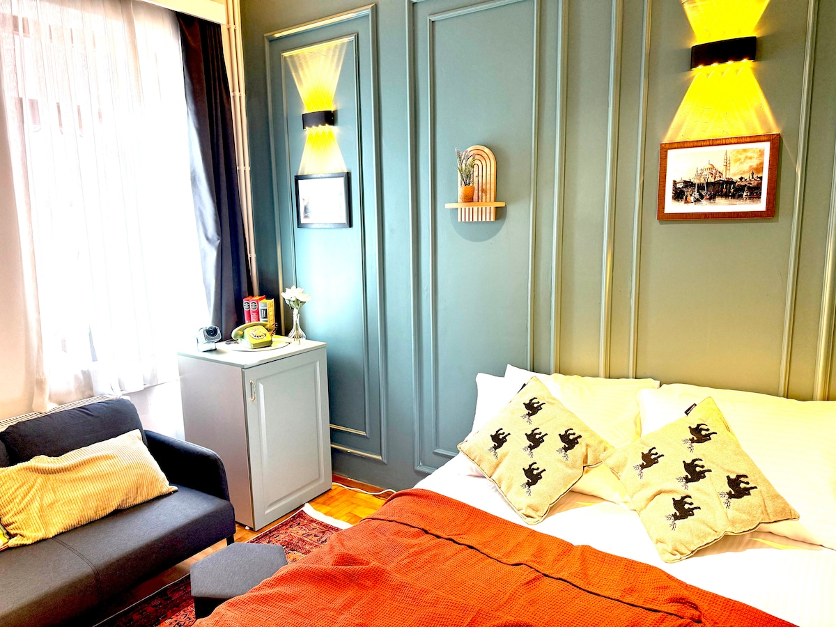 A cozy bedroom features a spacious bed with patterned pillows. A soft orange blanket is draped across the bed. A comfortable seating area with a dark sofa is positioned adjacent to a light-colored cabinet. Warm lighting accents the textured blue walls, creating a soothing ambiance.