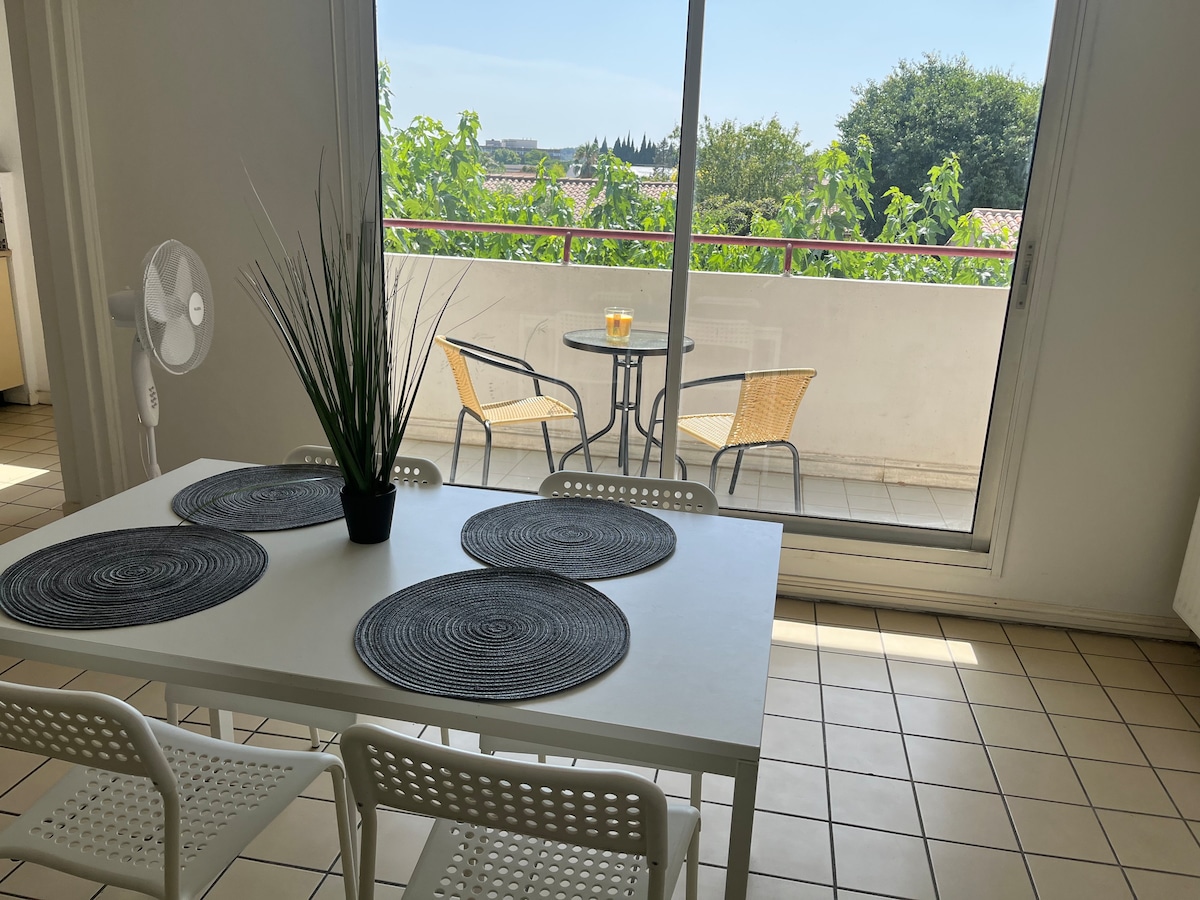 A dining area features a white table with four chairs and decorative placemats. Large sliding glass doors provide access to a balcony, where a small round table and two chairs are positioned. Natural light floods the space, highlighting the surrounding greenery.