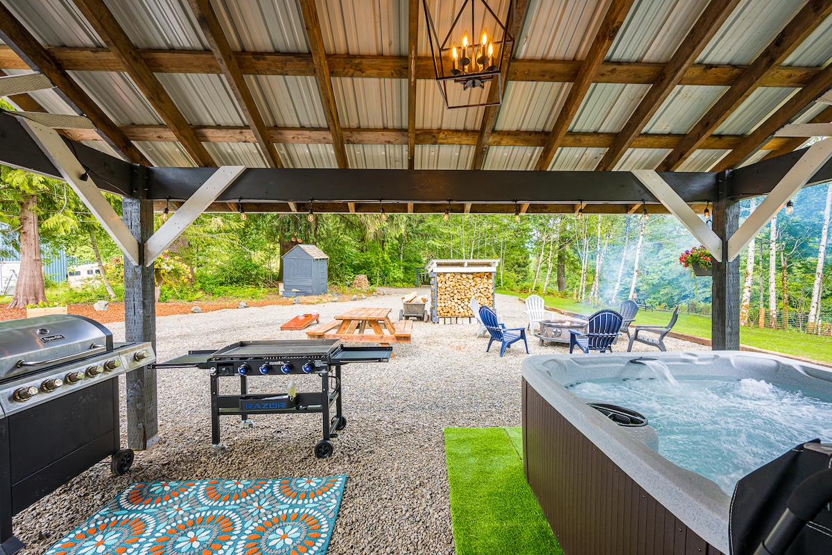 An outdoor entertainment area is visible, featuring a covered space with a hot tub, a grill, and a dining grill. Surrounding the area, seating is arranged with blue Adirondack chairs and a fire pit with wooden benches. A gravel path is seen leading to additional landscaping.