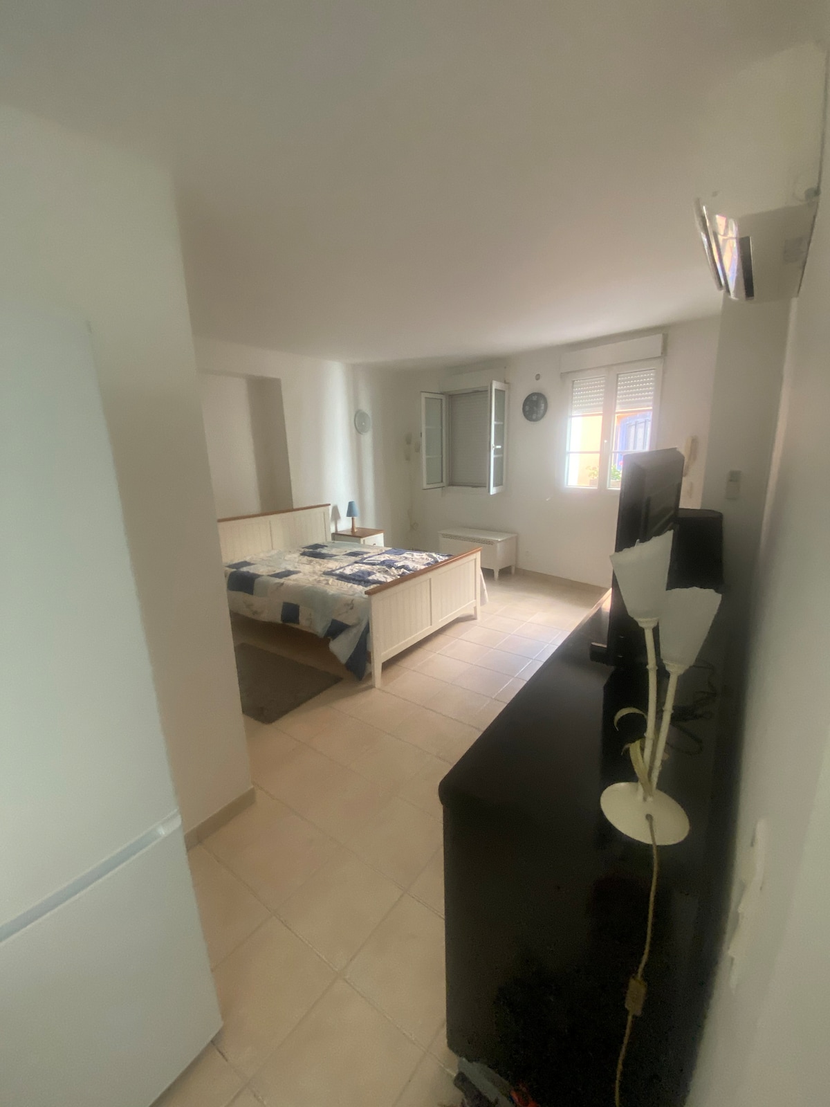 A neat studio space is seen, featuring a spacious bed adorned with a patterned blanket. A mirrored wardrobe and a television are positioned against a light-colored wall. Natural light enters through a window, illuminating the shimmery tiles of the floor.