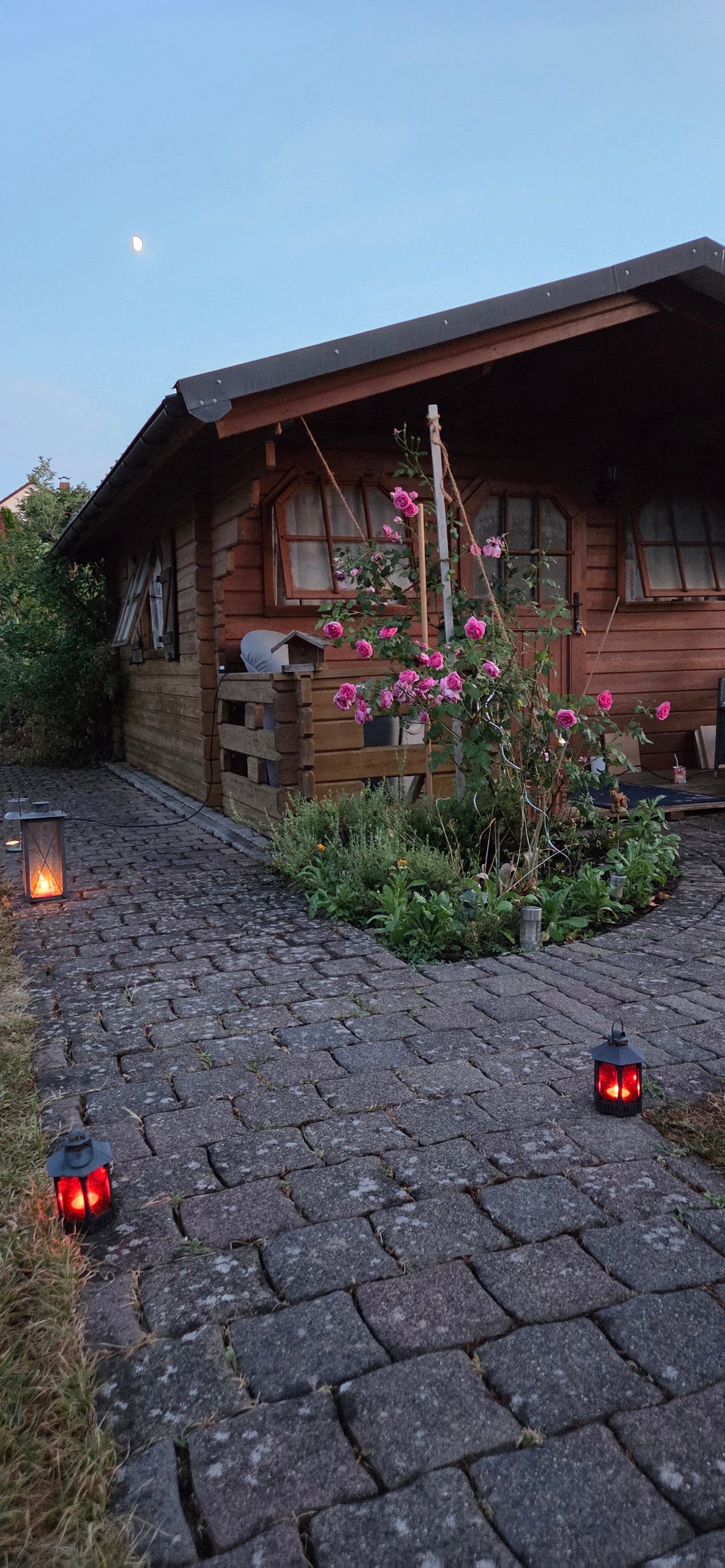 A wooden chalet is framed by lush greenery and vibrant flowers. A stone pathway leads to the entrance, illuminated by lanterns that cast a soft glow. The sky above features a hint of twilight, suggesting a peaceful evening ambiance.