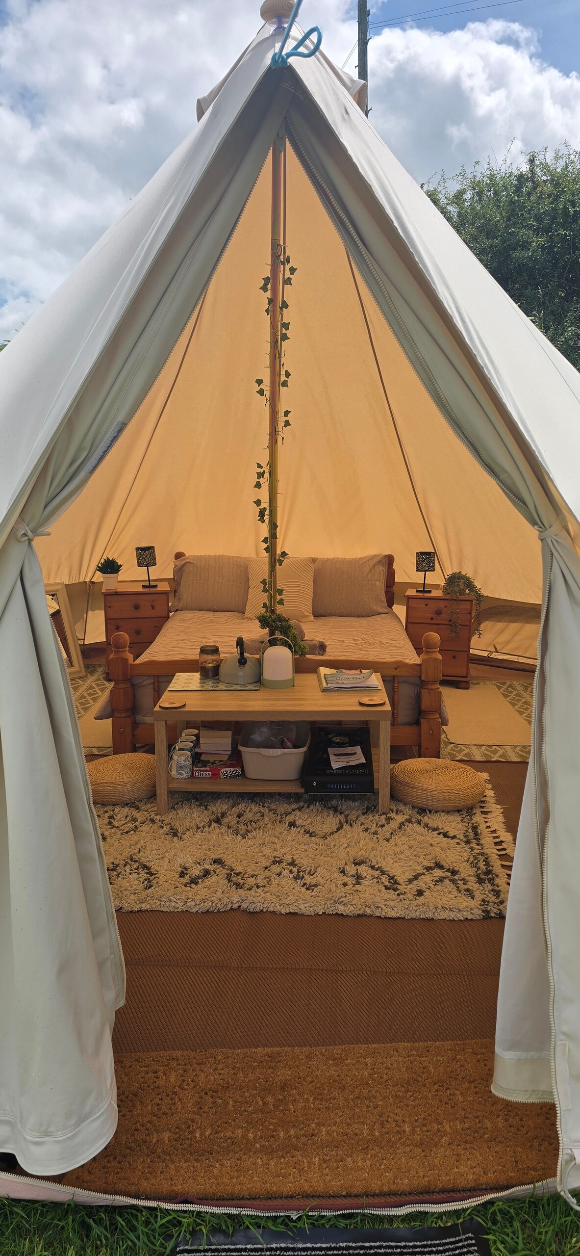 The interior of the bell tent is warmly lit, featuring a central wooden table surrounded by comfortable seating. Two bedside tables with lamps are positioned alongside a spacious area, adorned with rugs and cushions, creating a cozy atmosphere for relaxation.