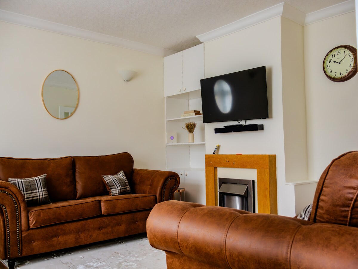 A living room is presented with two brown leather sofas, each featuring plaid accent pillows. A modern flat-screen TV is mounted above a simple fireplace on a light-colored wall. A round mirror and a clock accentuate the space, providing a clean and cozy atmosphere.