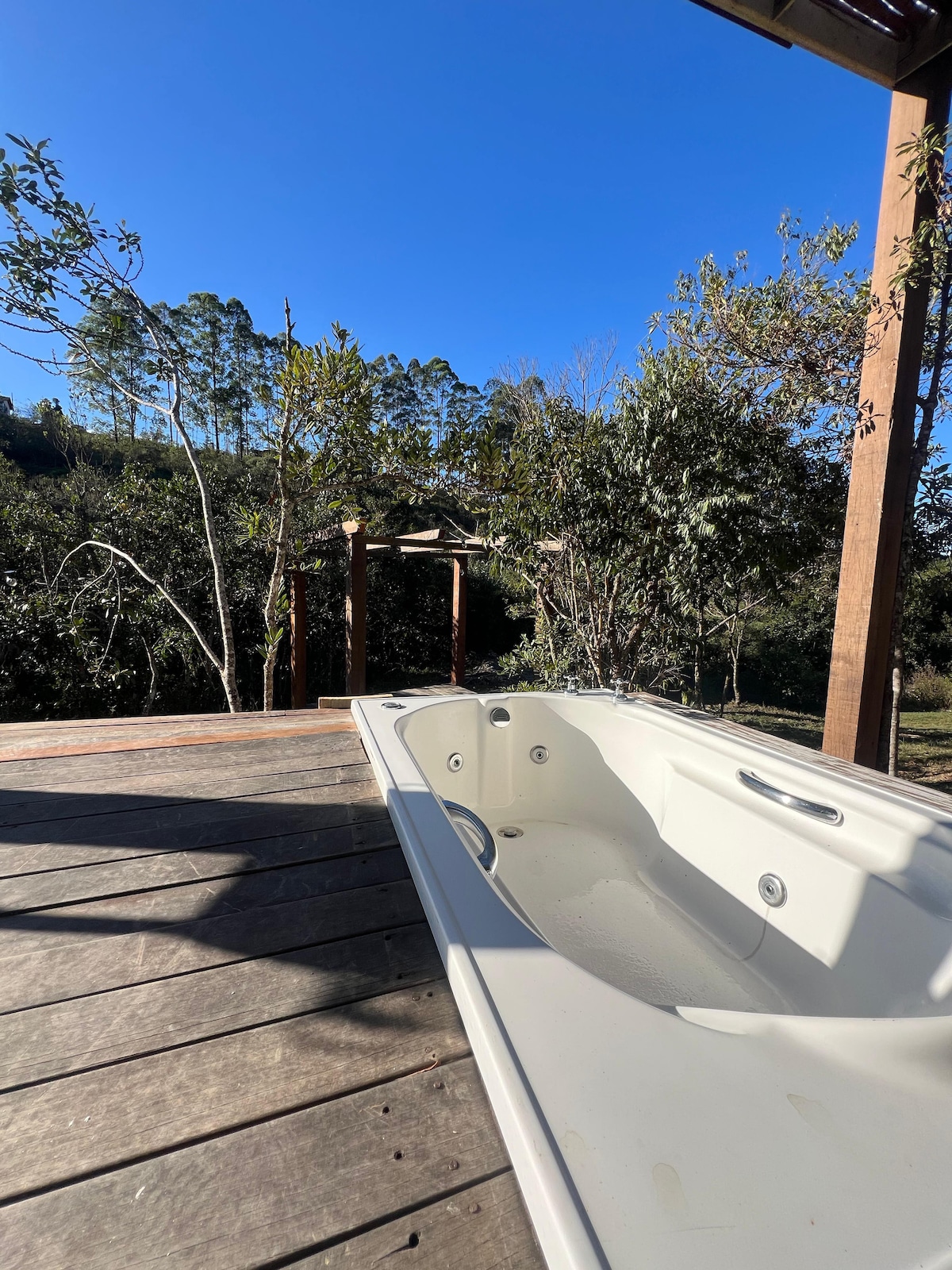 A private outdoor area features a spacious hot tub situated on a wooden deck. Surrounding greenery, including trees and shrubs, provide a natural backdrop, with clear blue sky visible overhead.