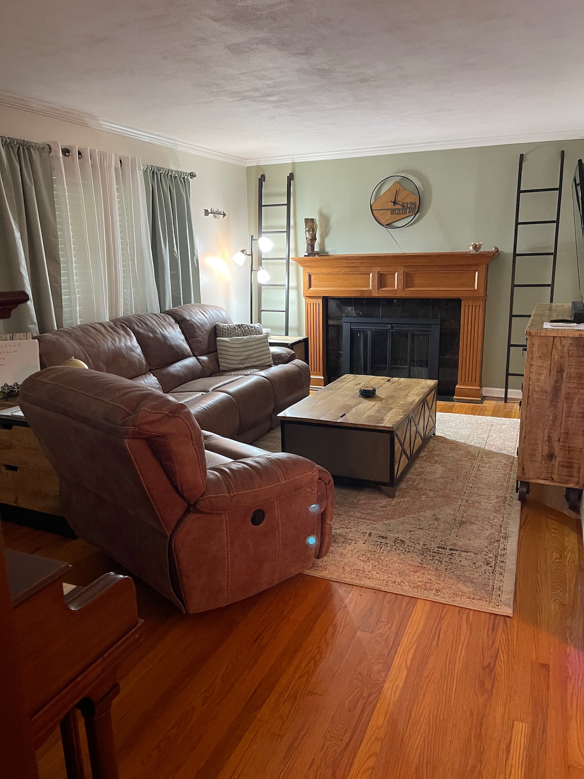 A comfortable living room is featured with a brown sectional sofa facing a wooden coffee table. A fireplace with a mantel is set against a pale green wall, complemented by light-colored curtains and two metal ladders. Wooden flooring enhances the space.