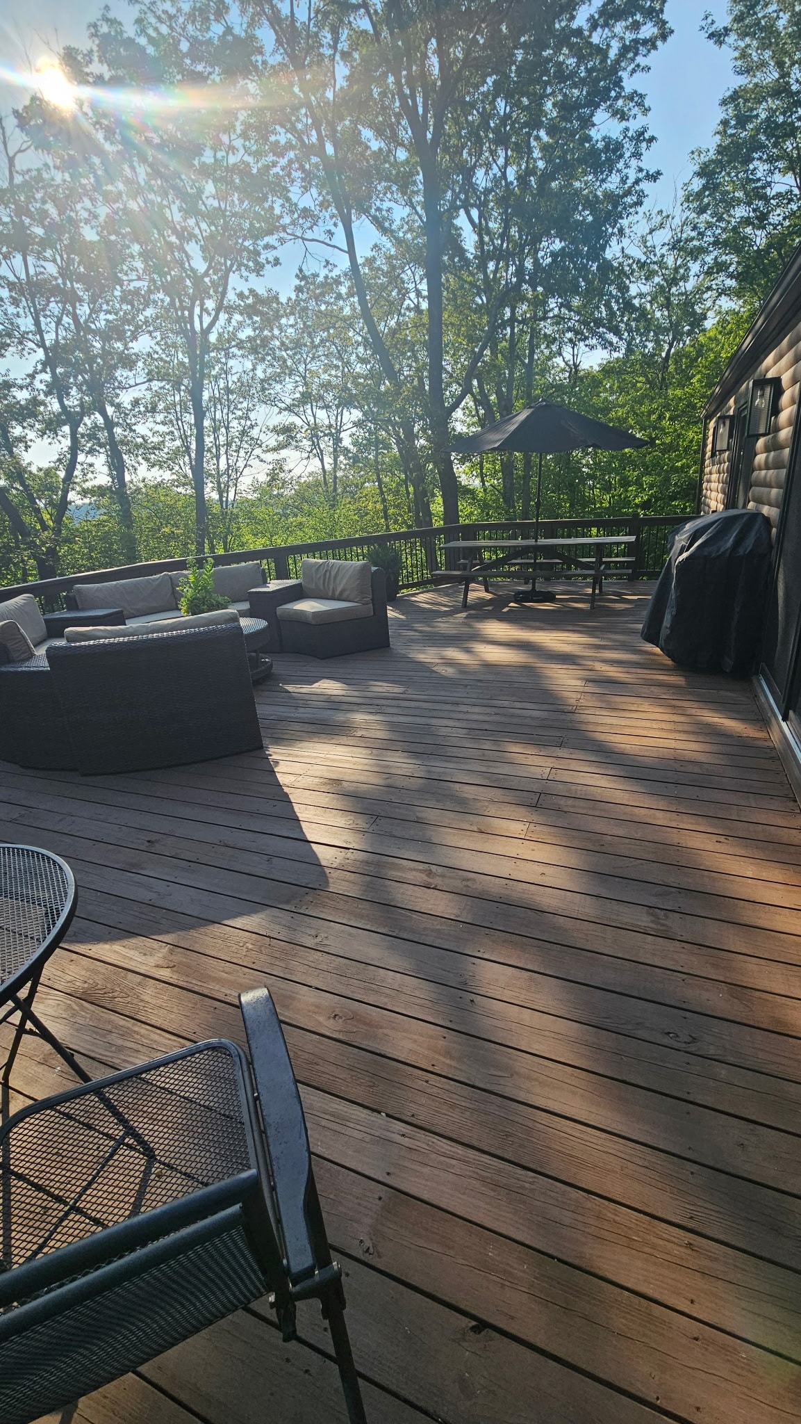 A spacious wooden deck is illuminated by sunlight, surrounded by trees. Seating areas with comfortable chairs and a table are available, along with a grill for outdoor cooking. An umbrella provides shade in part of the seating area, enhancing the inviting atmosphere for relaxation.