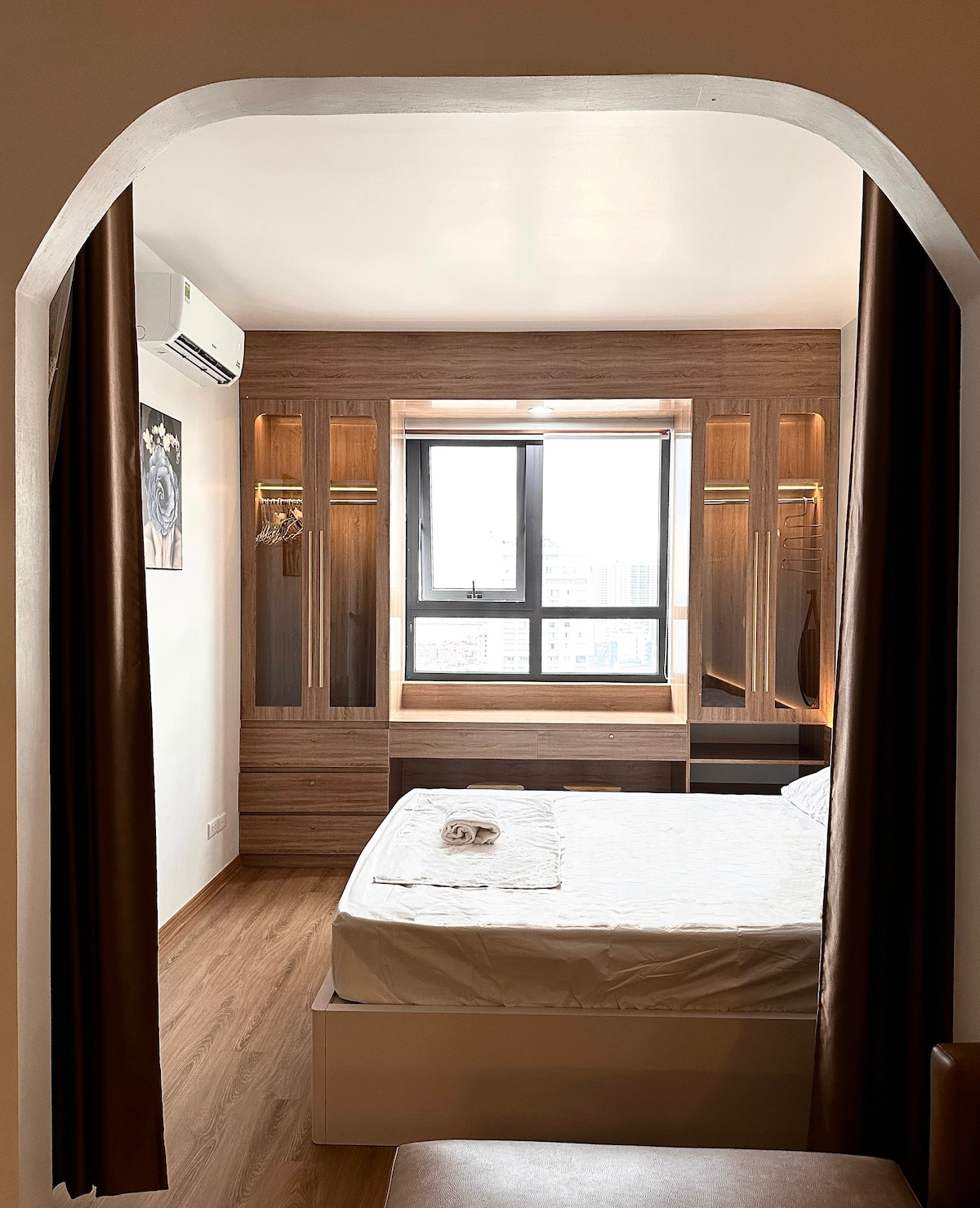 A well-lit sleeping area is highlighted by a spacious bed, accompanied by a neatly rolled towel. A large window provides city views and natural light, while the wooden cabinetry and soft lighting create a functional space. Dark curtains frame the window, enhancing privacy.