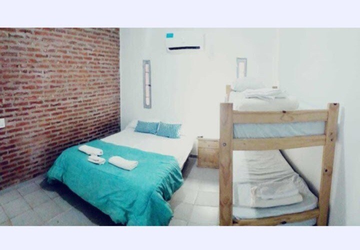 A bright sleeping area features a spacious bed with a soft blue blanket and two pillows. A wooden bunk bed stands adjacent, with neatly arranged bedding. Exposed brick walls add texture, while light fills the room through a window. Air conditioning is visible for comfort.