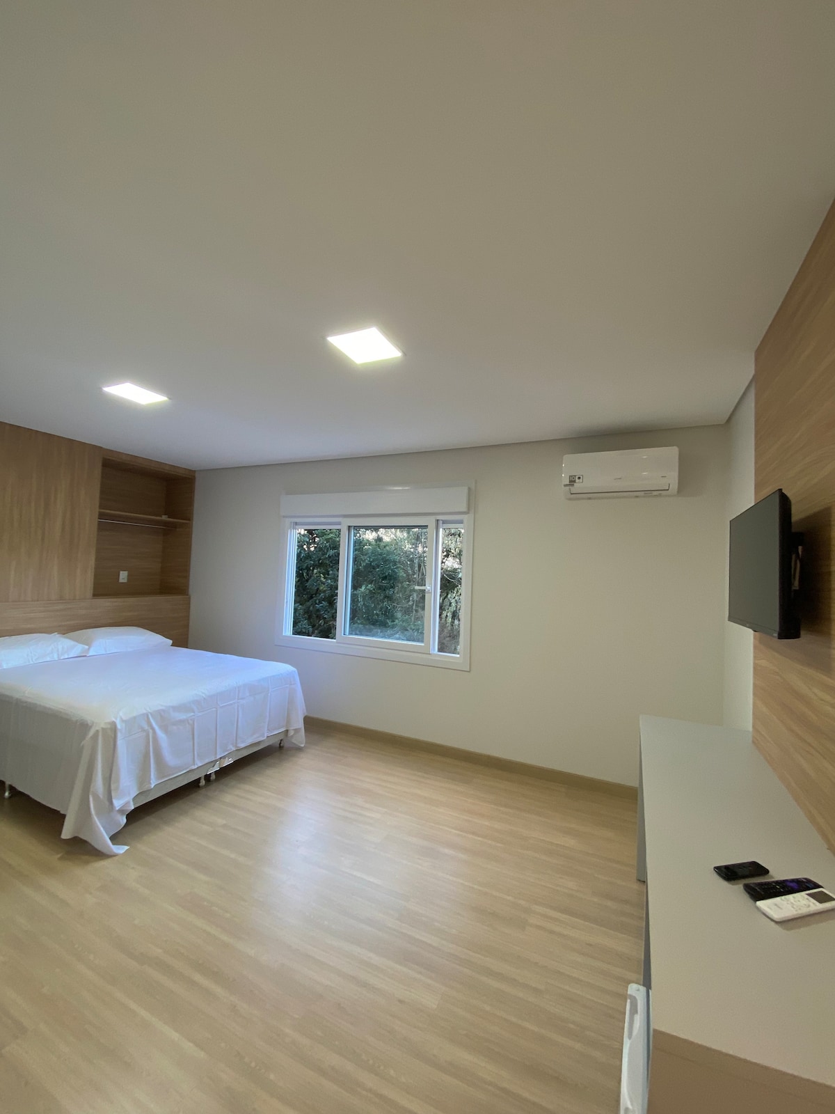 A spacious room is equipped with a comfortable bed covered with a light-colored blanket. Natural light enters through a large window, while modern lighting illuminates the space. An air conditioning unit and a television mounted on the wall enhance convenience.