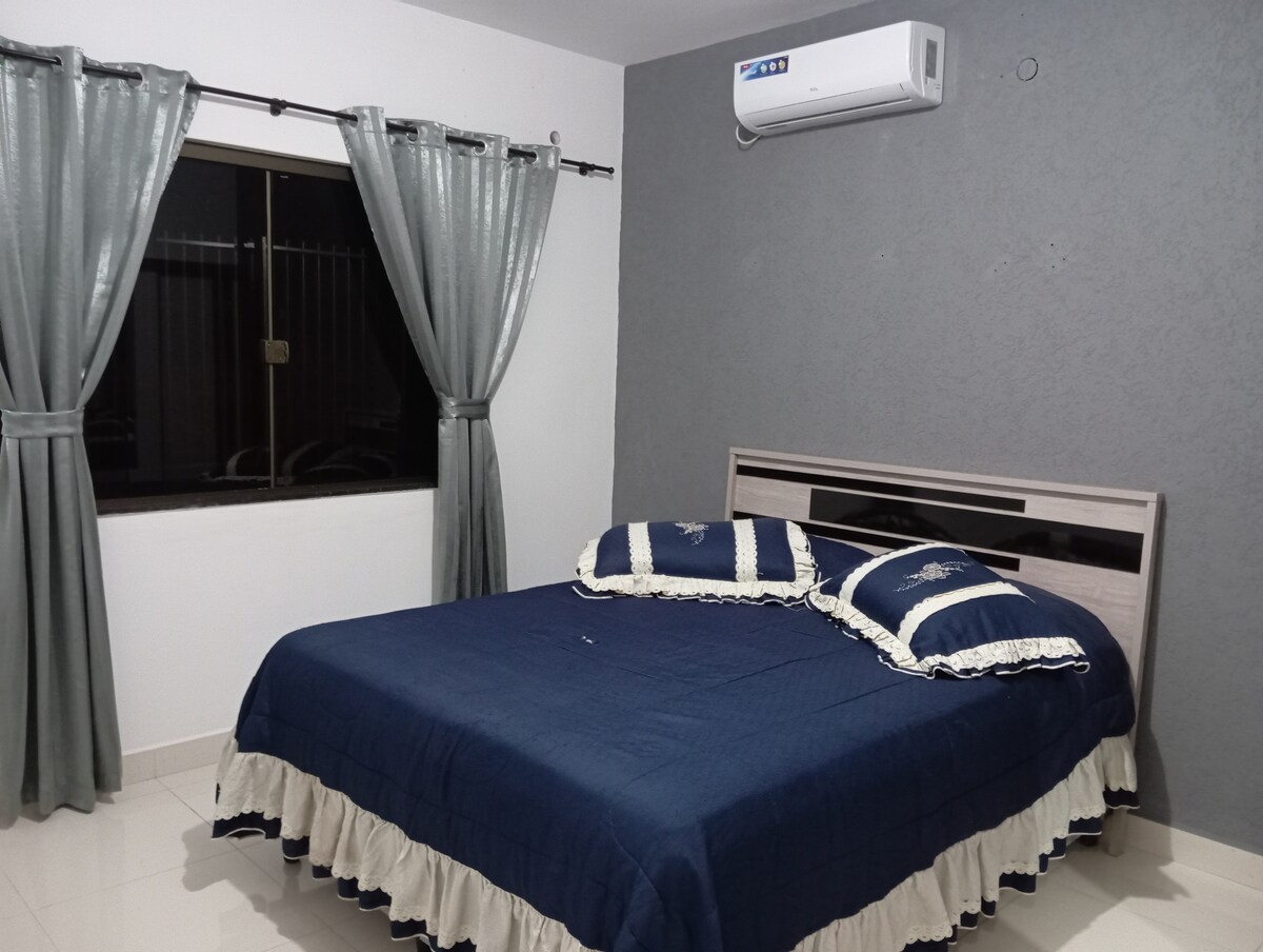 A sleeping area features a spacious bed with a dark blue coverlet and matching pillows. The room is illuminated by a nearby window, which is adorned with sheer gray curtains. An air conditioning unit is mounted above the bed, providing a cool atmosphere.