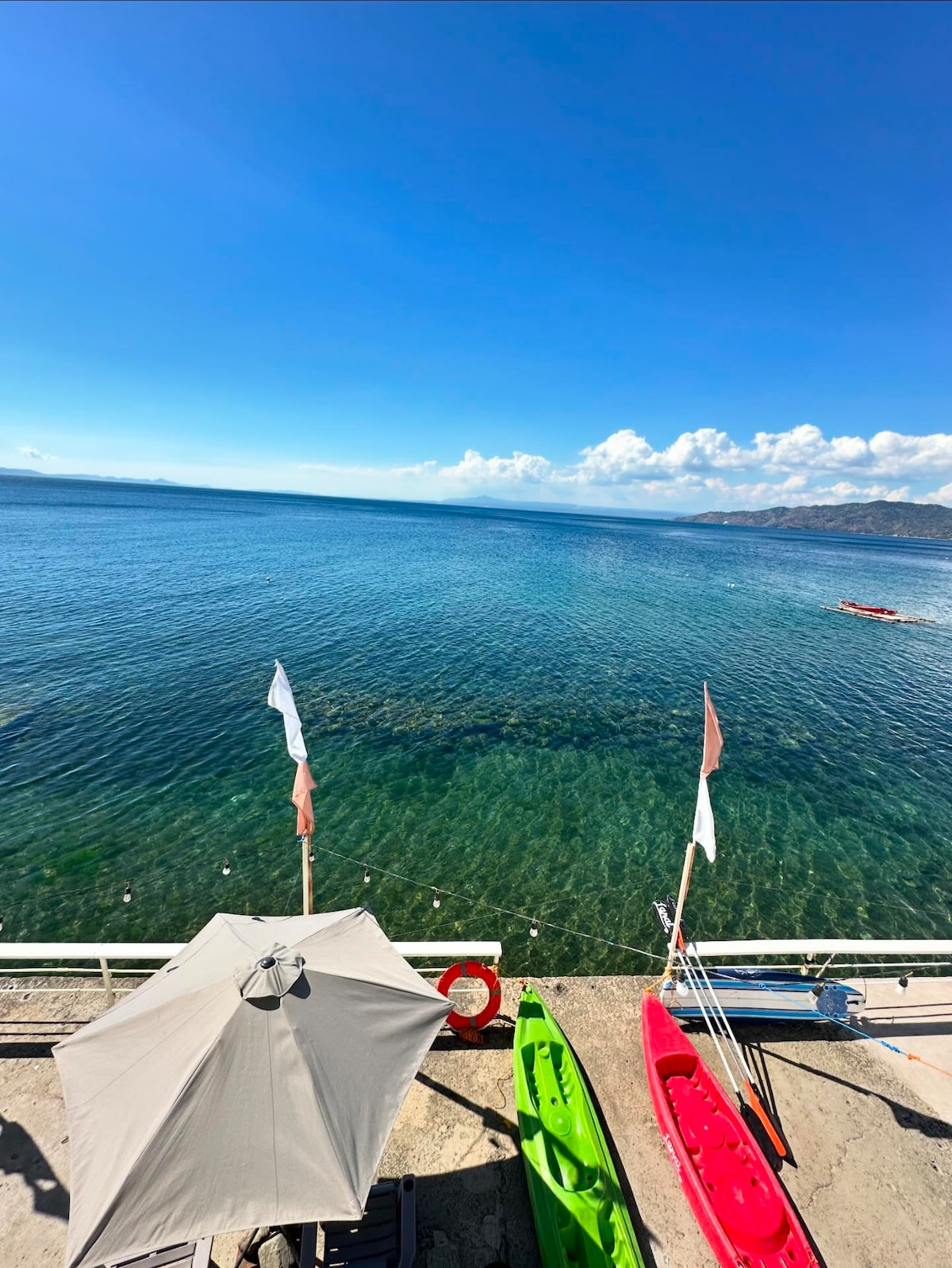 A vibrant view of a clear ocean stretches towards the horizon, complemented by a bright blue sky with a few scattered clouds. Colorful kayaks in green and red sit on a concrete area, while a sunshade provides a spot for relaxation near the water's edge.